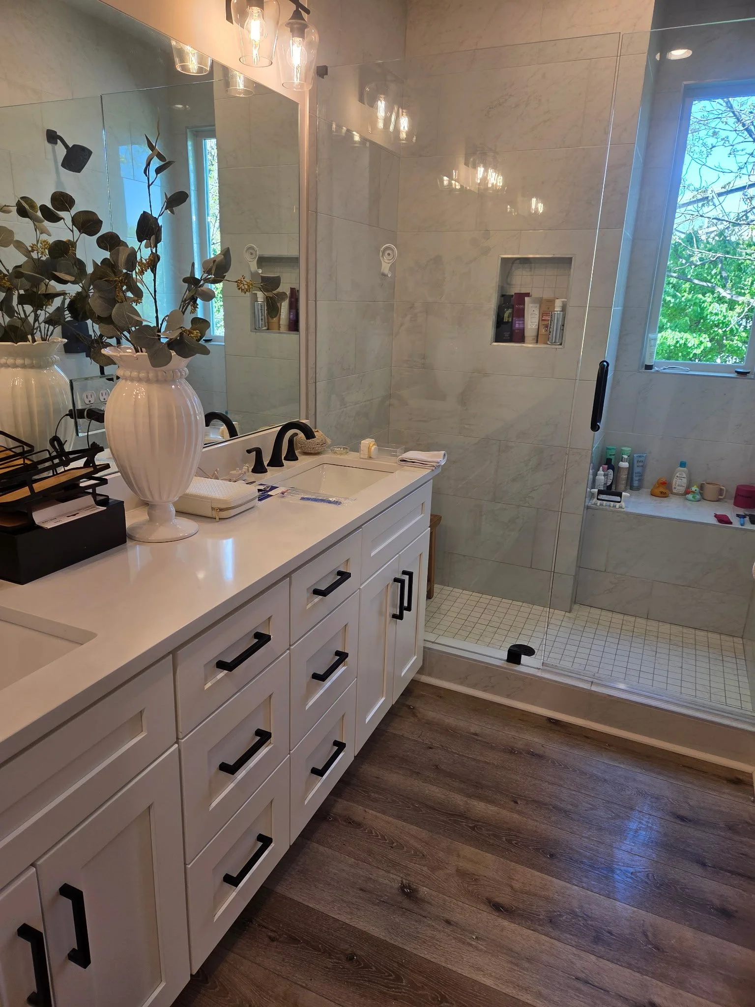 Bathroom with white vanity, black handles, large mirror, decorative vase with greenery, and a walk-in shower with a glass door, niche shelves with toiletries, and a window showing trees outside.