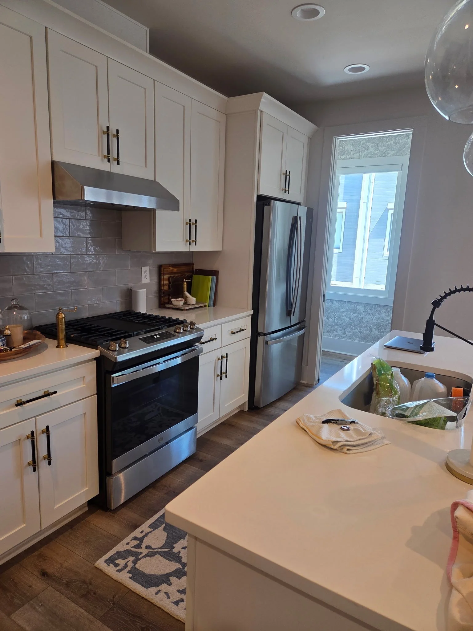 A modern kitchen with white cabinets, stainless steel refrigerator, gas stove, and a white countertop with a sink. There is a window in the background, and various kitchen items on the counter, including a chopping board, bottles, and a plastic bag with groceries. A patterned rug is on the wooden floor.
