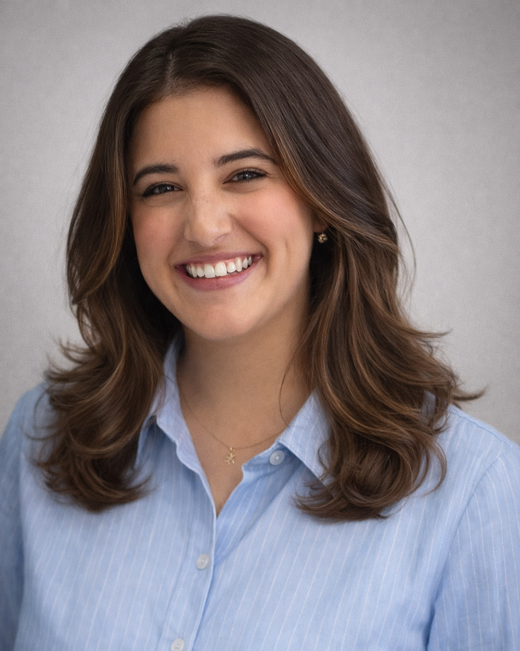 A young woman with shoulder-length brown hair and a warm smile, wearing a light blue button-up shirt, a small necklace, and earrings.
