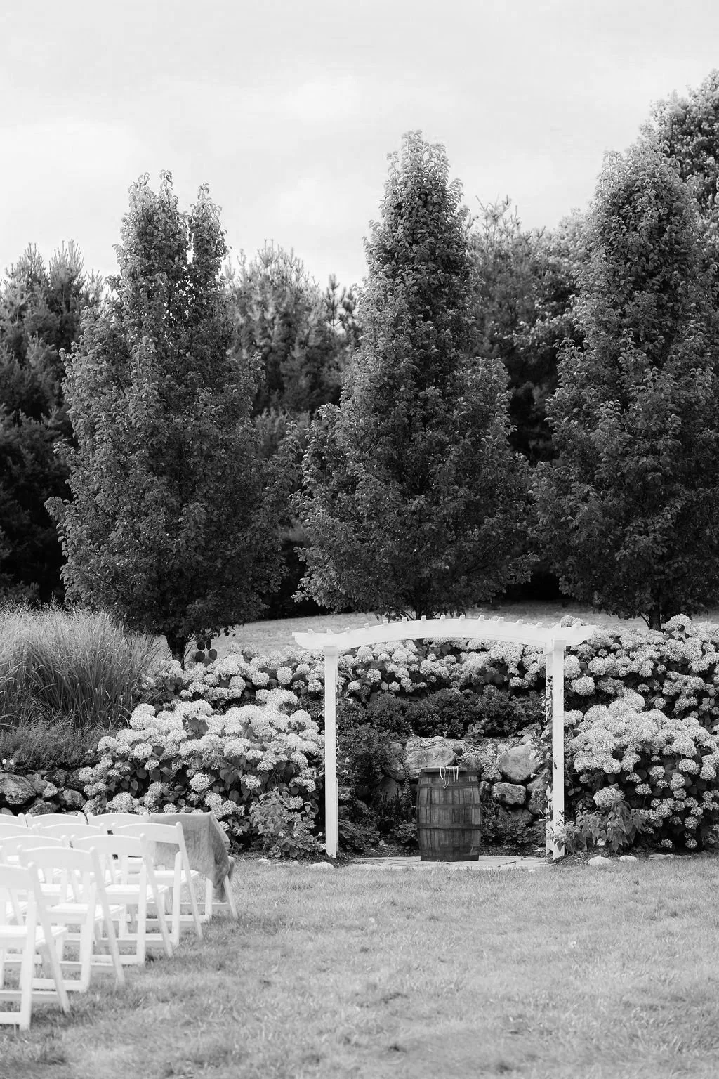 Outdoor wedding ceremony setup with white chairs arranged on grass, a decorative arch with white wood, and a barrel with flowers in the background, surrounded by trees and bushes.