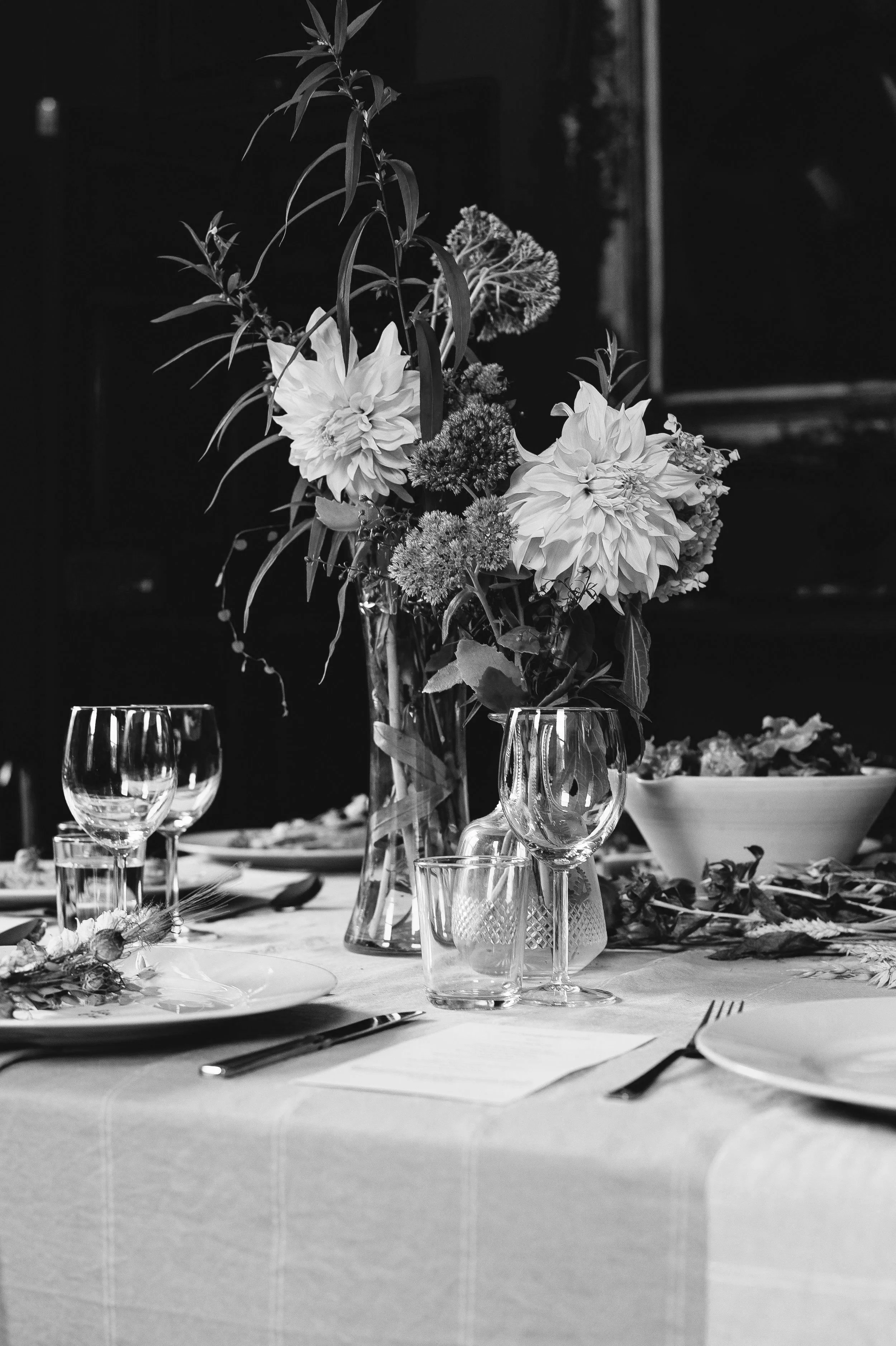 A black and white photo of a dining table decorated with a large floral centerpiece in a vase, surrounded by wine glasses, a bowl, and plates.