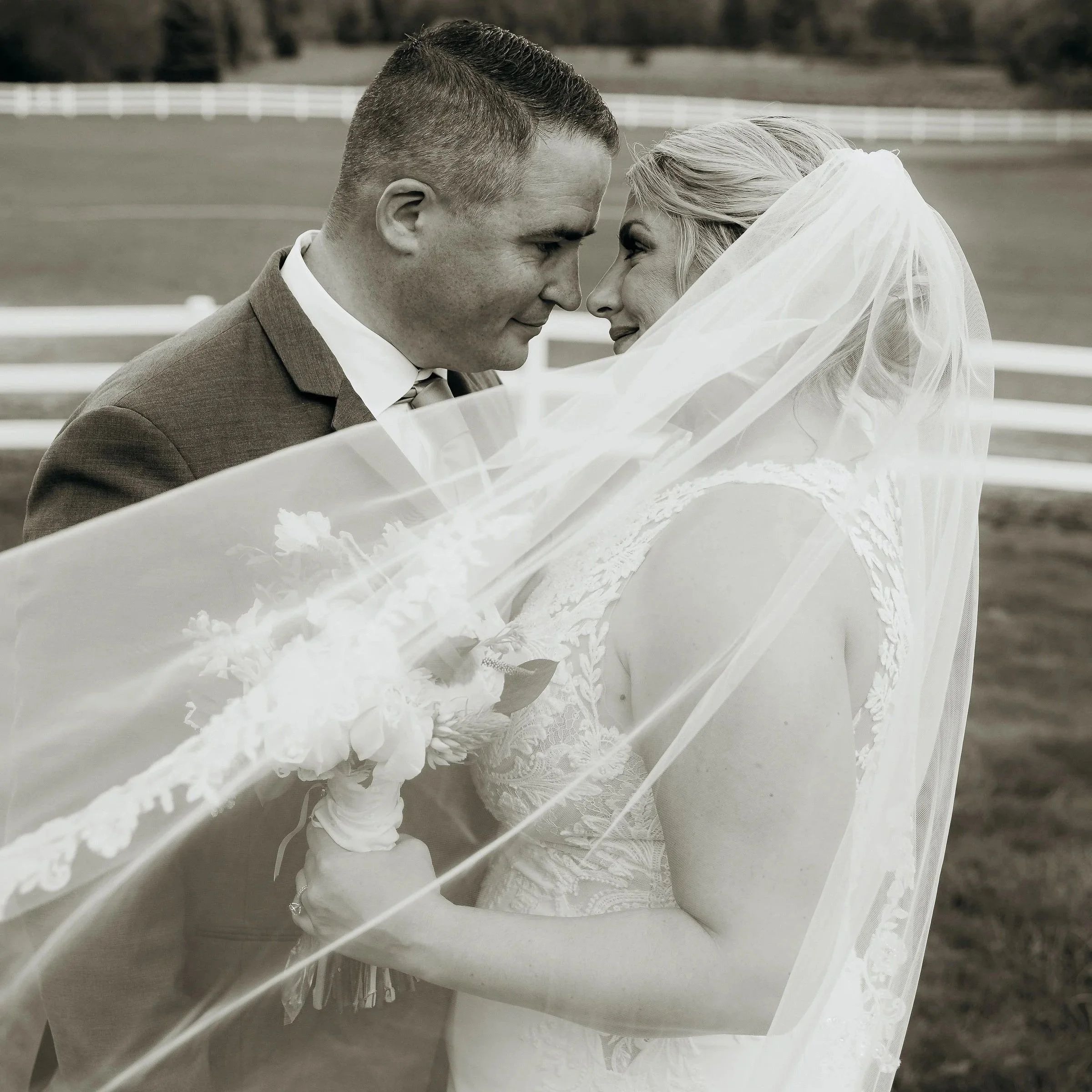 Black and white photo of a bride and groom touching foreheads under the bride's veil, outdoors on a grassy field with a white fence in the background, the bride holding a bouquet.