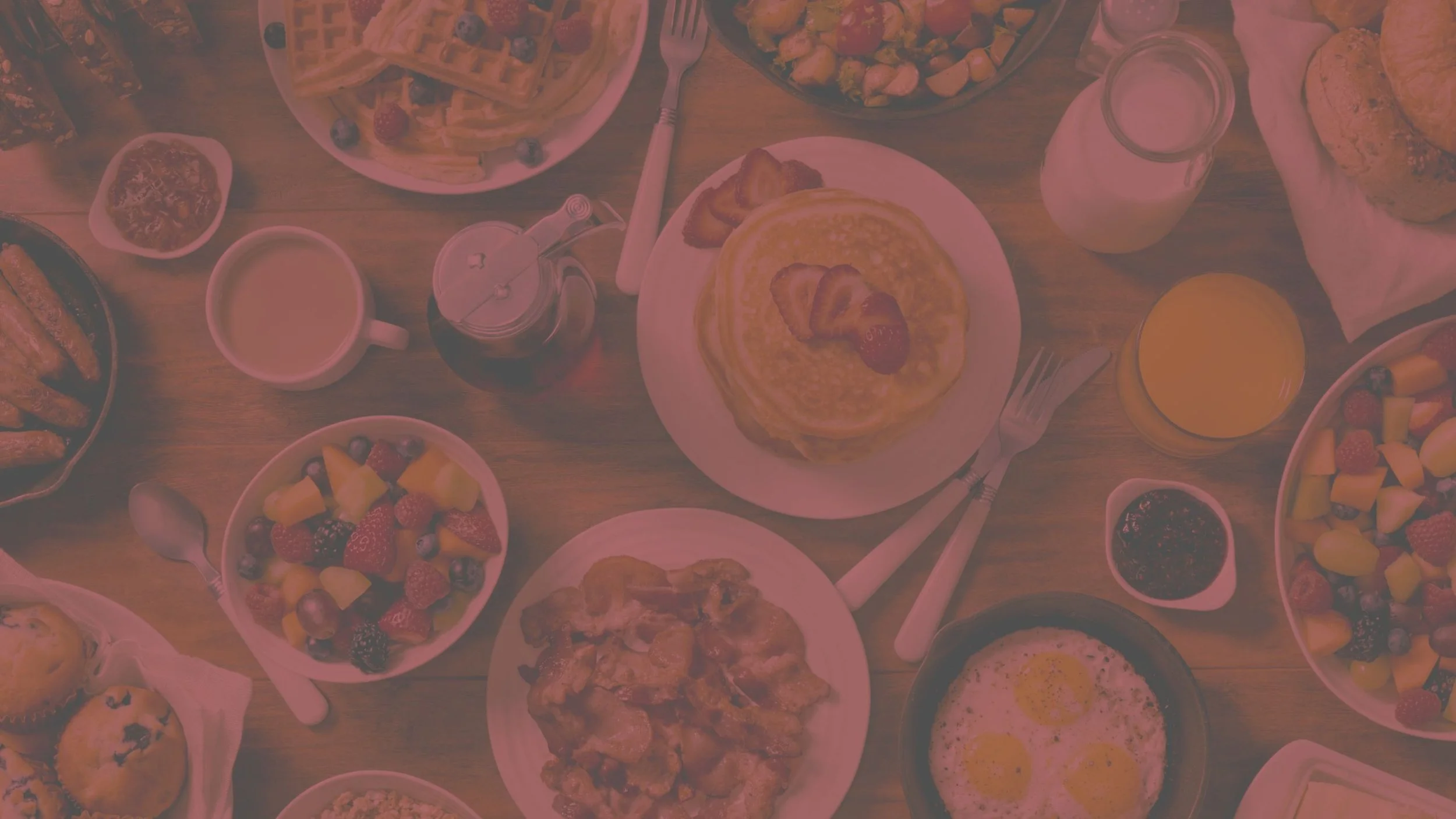 A breakfast table filled with various foods including waffles with strawberries, scrambled eggs, fruit bowls with berries and pineapple, bacon, muffins, toast, a glass of orange juice, a glass of milk, coffee, and a small bowl of jam.