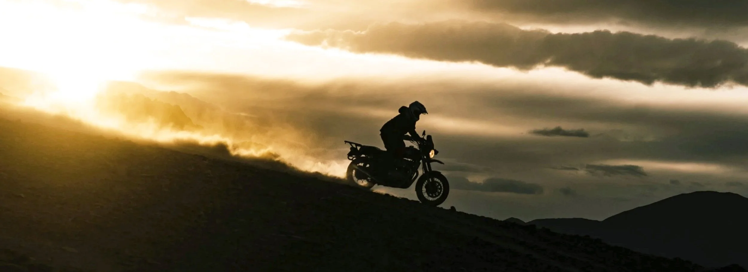 Silhouette of a person riding a motorcycle on a hill at sunset or sunrise with dramatic clouds in the sky.