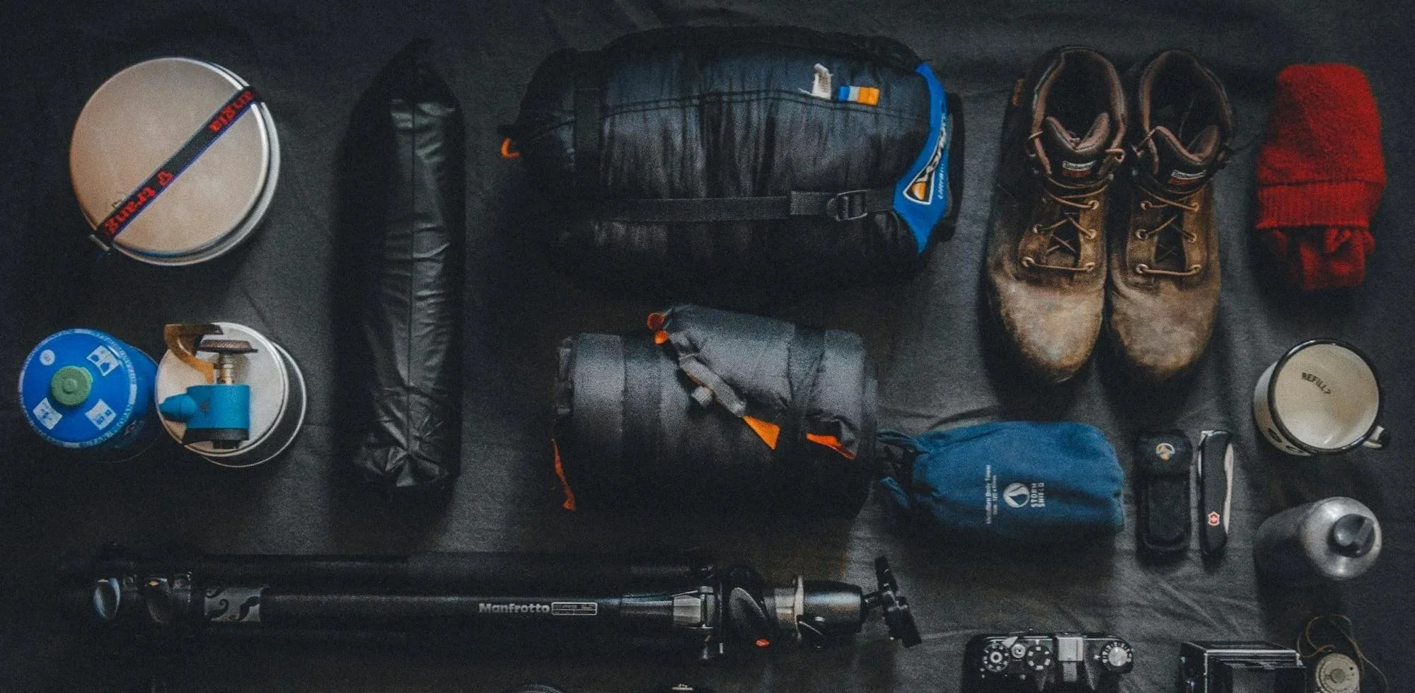 Camping or hiking gear laid out on a black surface, including a tripod, two pots, a gas canister, a small blue bag, a larger black bag, sleeping bags, hiking boots, a red knit hat, a cup, a water bottle, a Swiss Army knife, a camera, and other accessories.