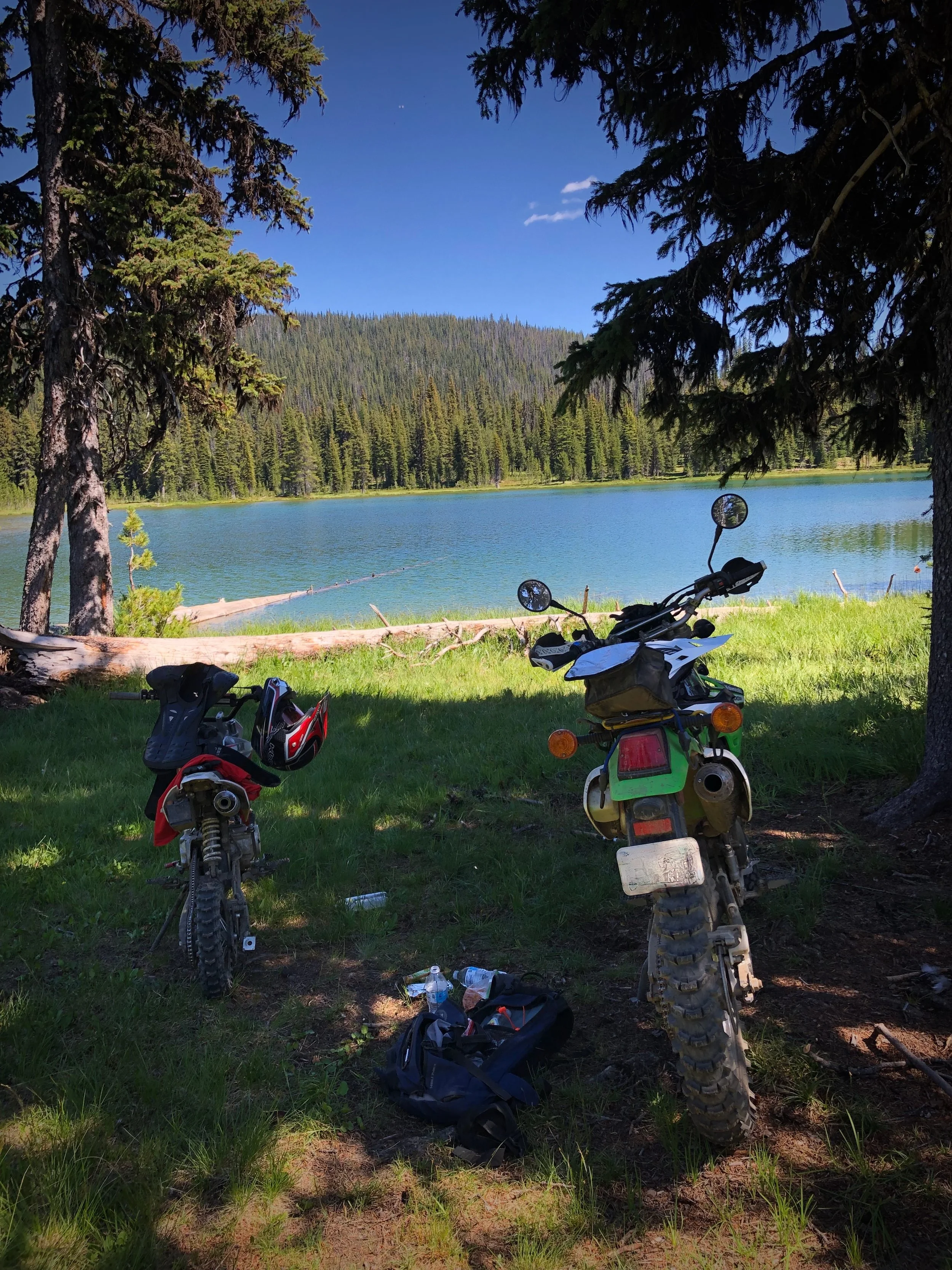 Two dirt bikes parked on a grassy area near a lake, with trees and pine forest in the background under a blue sky.