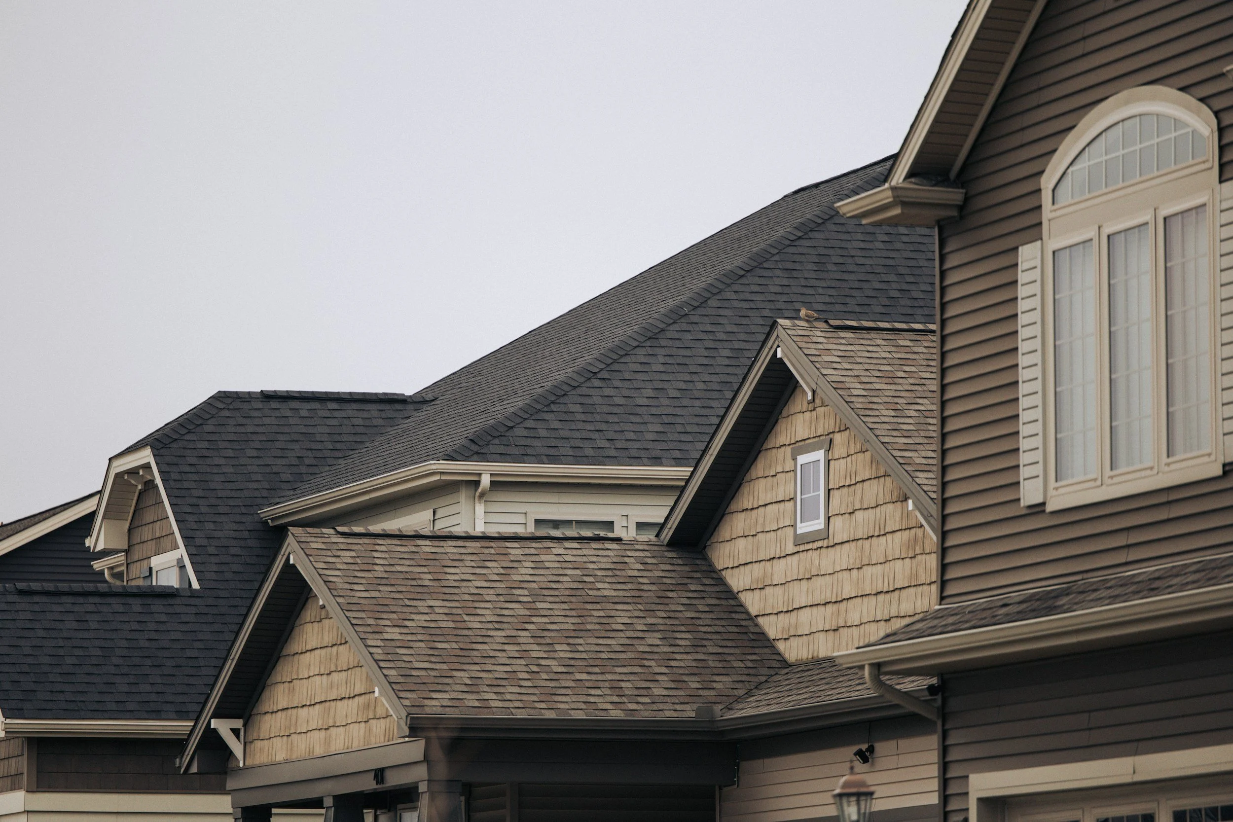 Close-up view of the rooftops of multiple residential houses with shingles, showing various angles and roof designs against a cloudy sky.