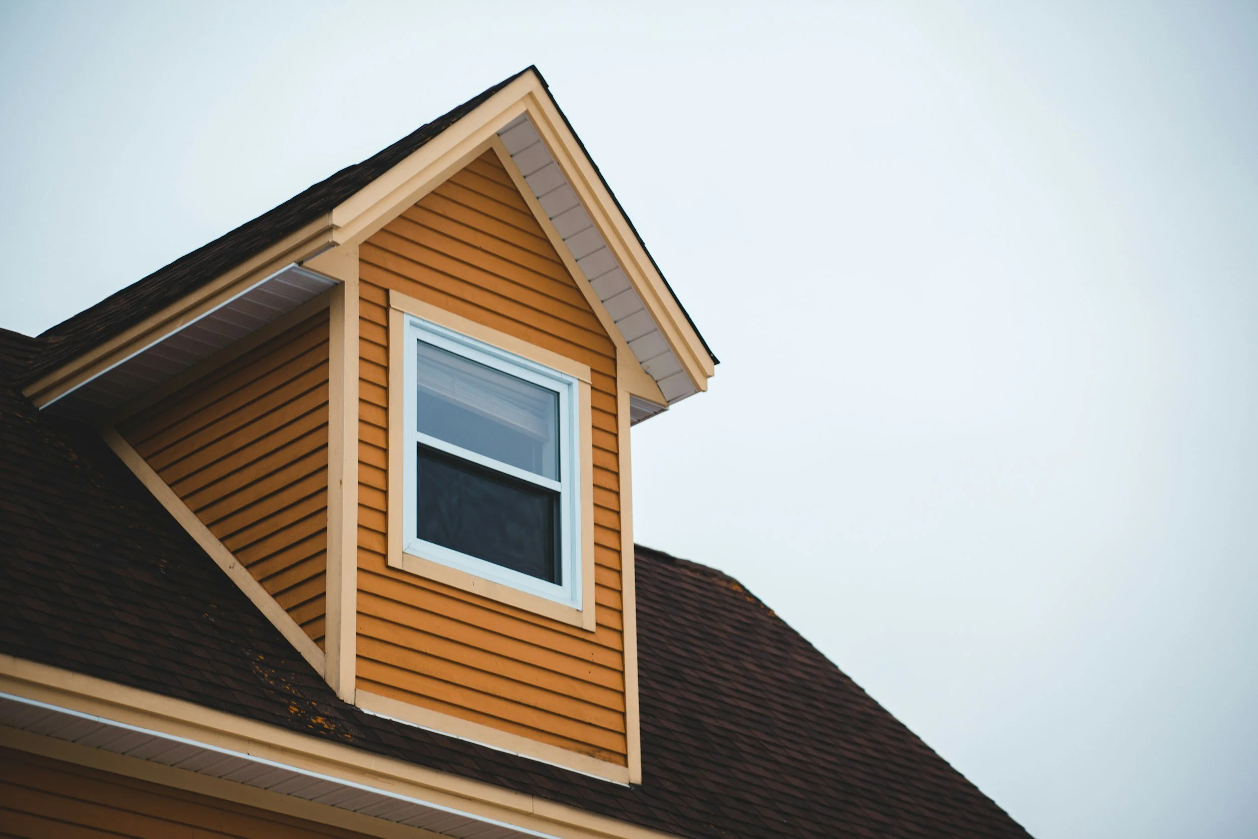 Close-up of a house's attic window with a brown gabled roof, orange siding, and white trim under a cloudy sky.