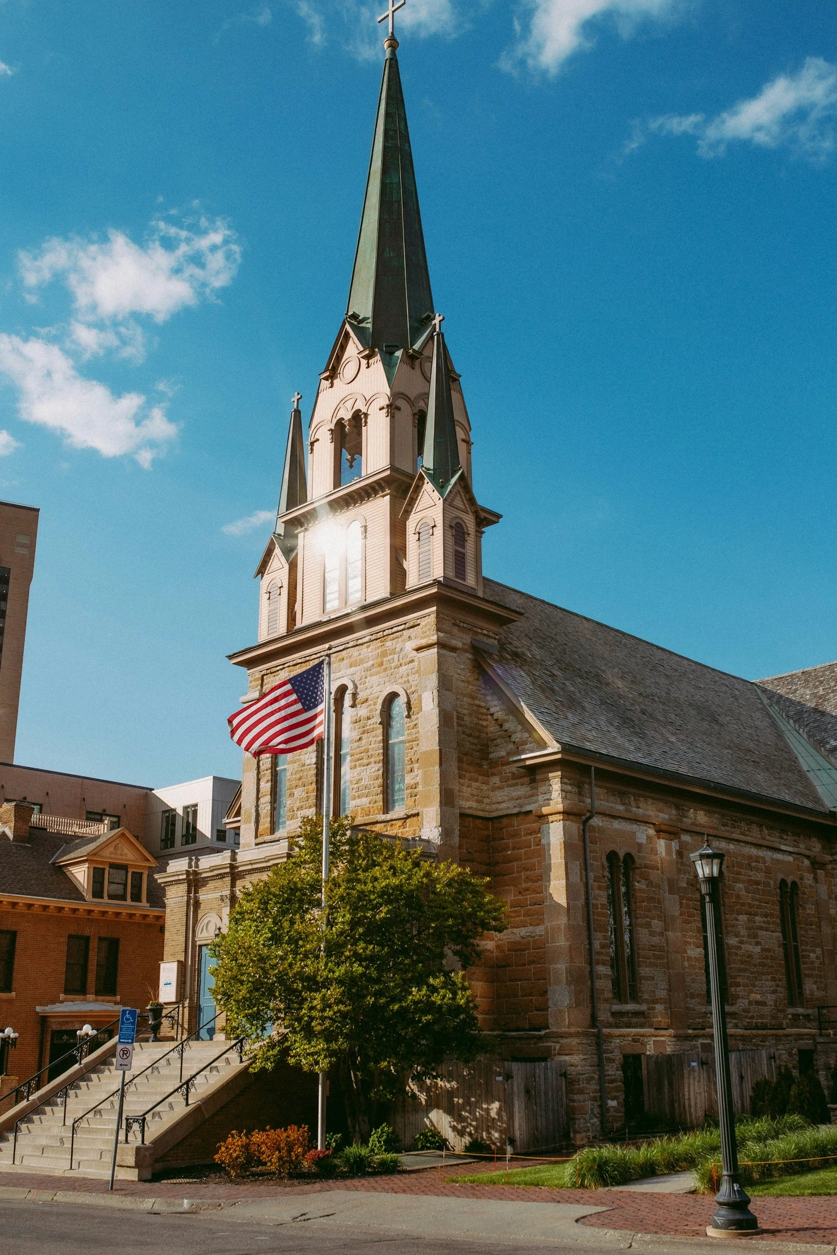 A stone church with a tall steeple topped with a cross, American flag on a pole in front, stairs leading to the entrance, and a tree with green foliage.