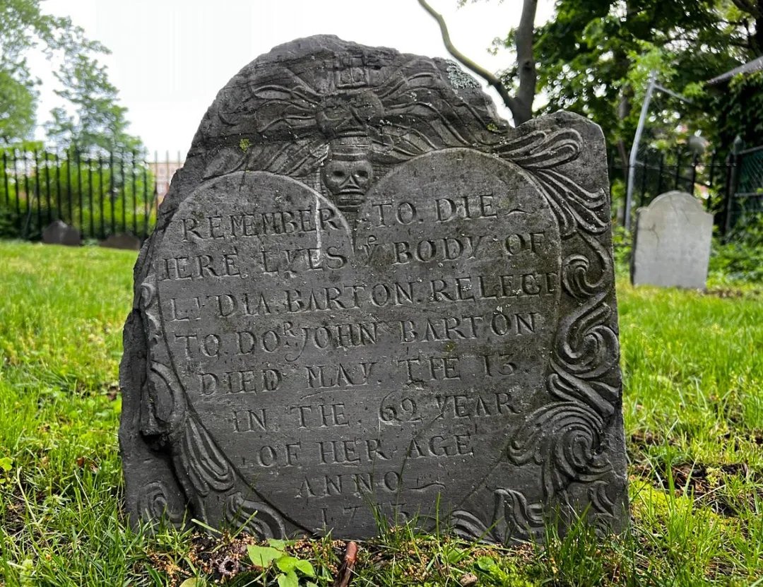 A weathered gravestone in a green cemetery, decorated with ornate carvings and a skull at the top, inscribed with a tribute to Ledda Barton, who died at age 69 in 1717.