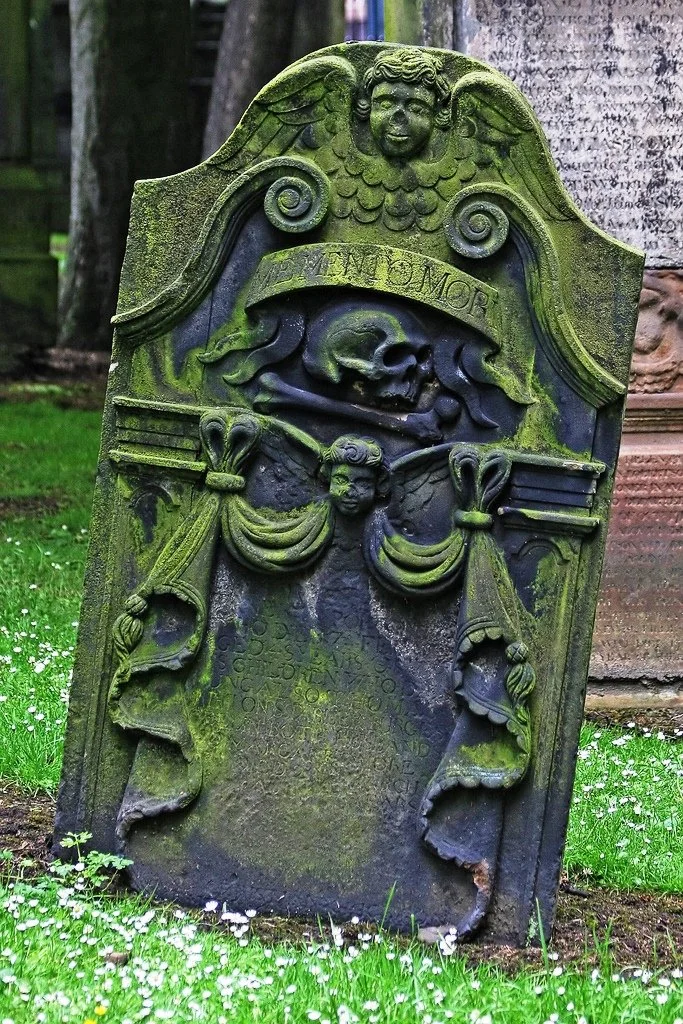 Old, moss-covered gravestone with ornate carvings including angel wings, a face at the top, and curtains draped across the middle, located in a graveyard with grass and small white flowers.
