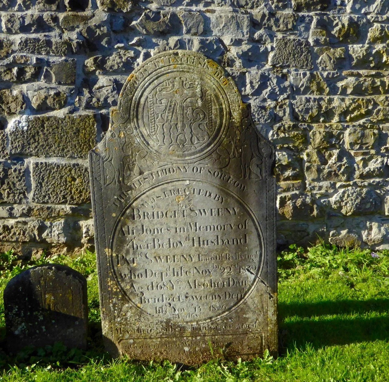 A historical tombstone made of gray stone, set against a stone wall, with moss and lichen. The inscription commemorates Bridget Sweeney, wife of John Sweeney, with details of her death, age, and her husband's memorial.