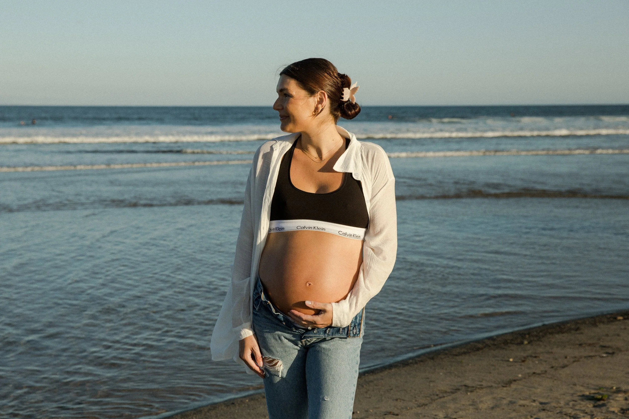 Pregnant woman standing on the beach near the shoreline, dressed in casual clothing and smiling, during sunset or late afternoon.