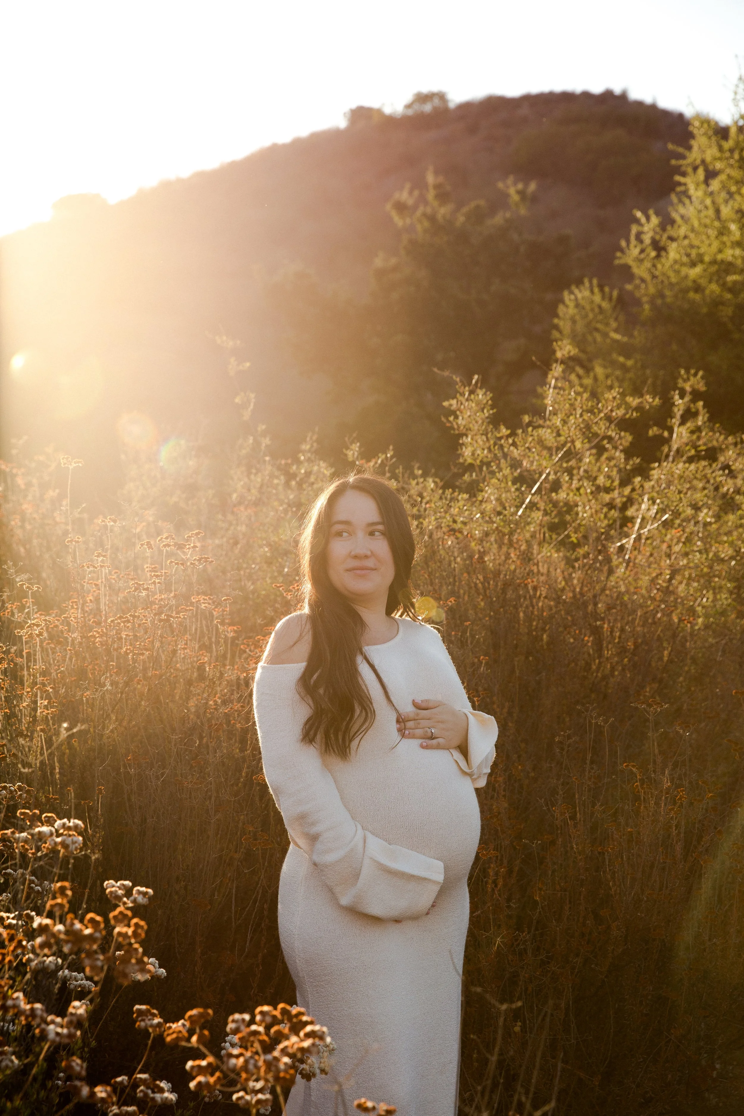 A pregnant woman with long dark hair standing outdoors in a field of wildflowers during sunset, wearing a white dress, with her hand on her belly, looking to the side.