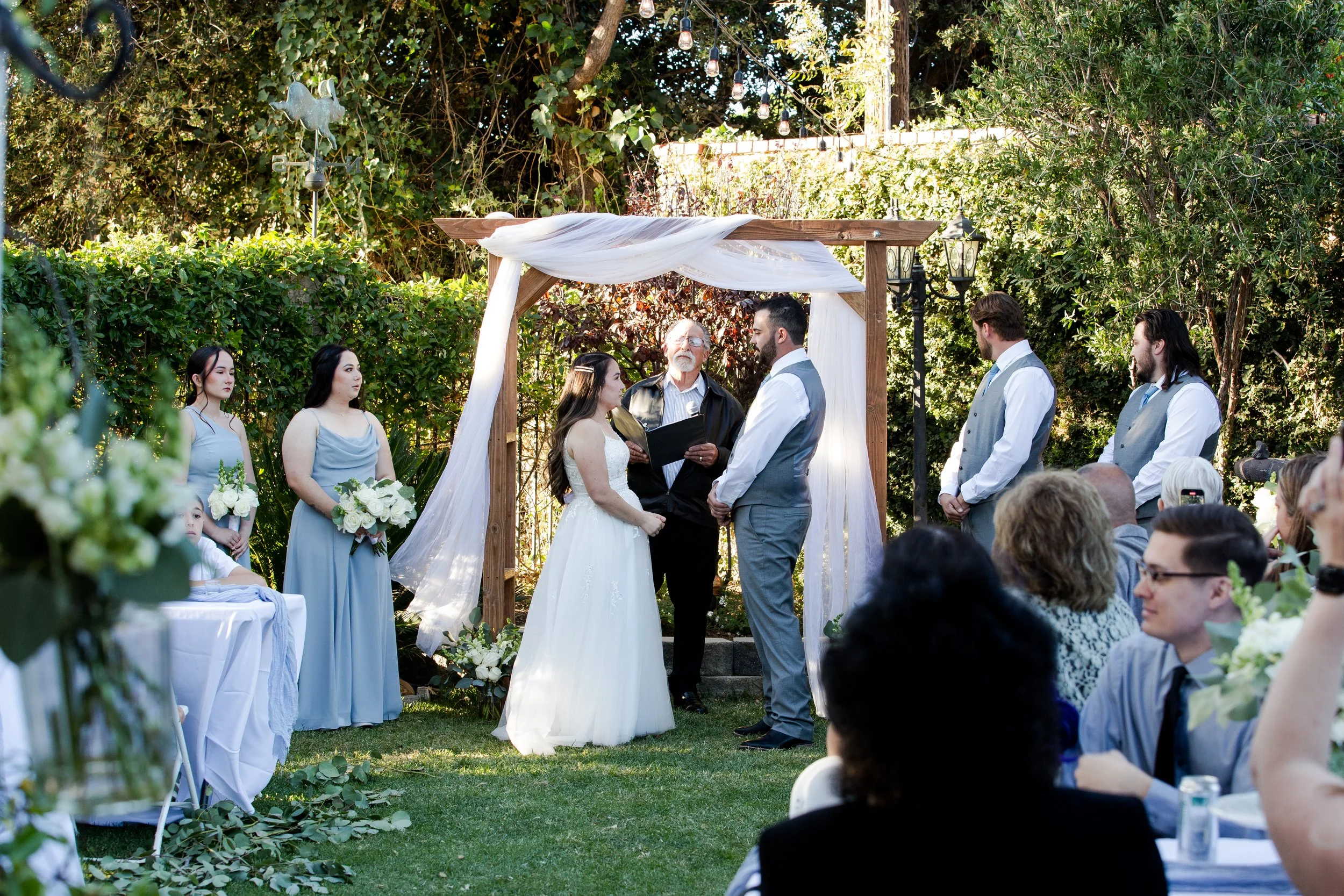 Beautiful outdoor wedding ceremony with a couple standing under a wooden arch draped with white fabric, surrounded by bridesmaids in light blue dresses and groomsmen in gray vests, and guests seated at tables decorated with white flowers.