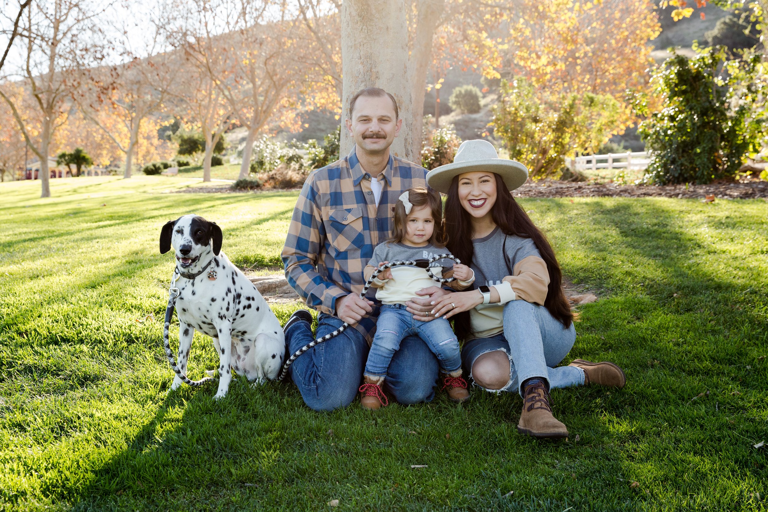 Family of three sitting on grass with their Dalmatian dog, autumn trees in the background, sunny day, smiling and happy.