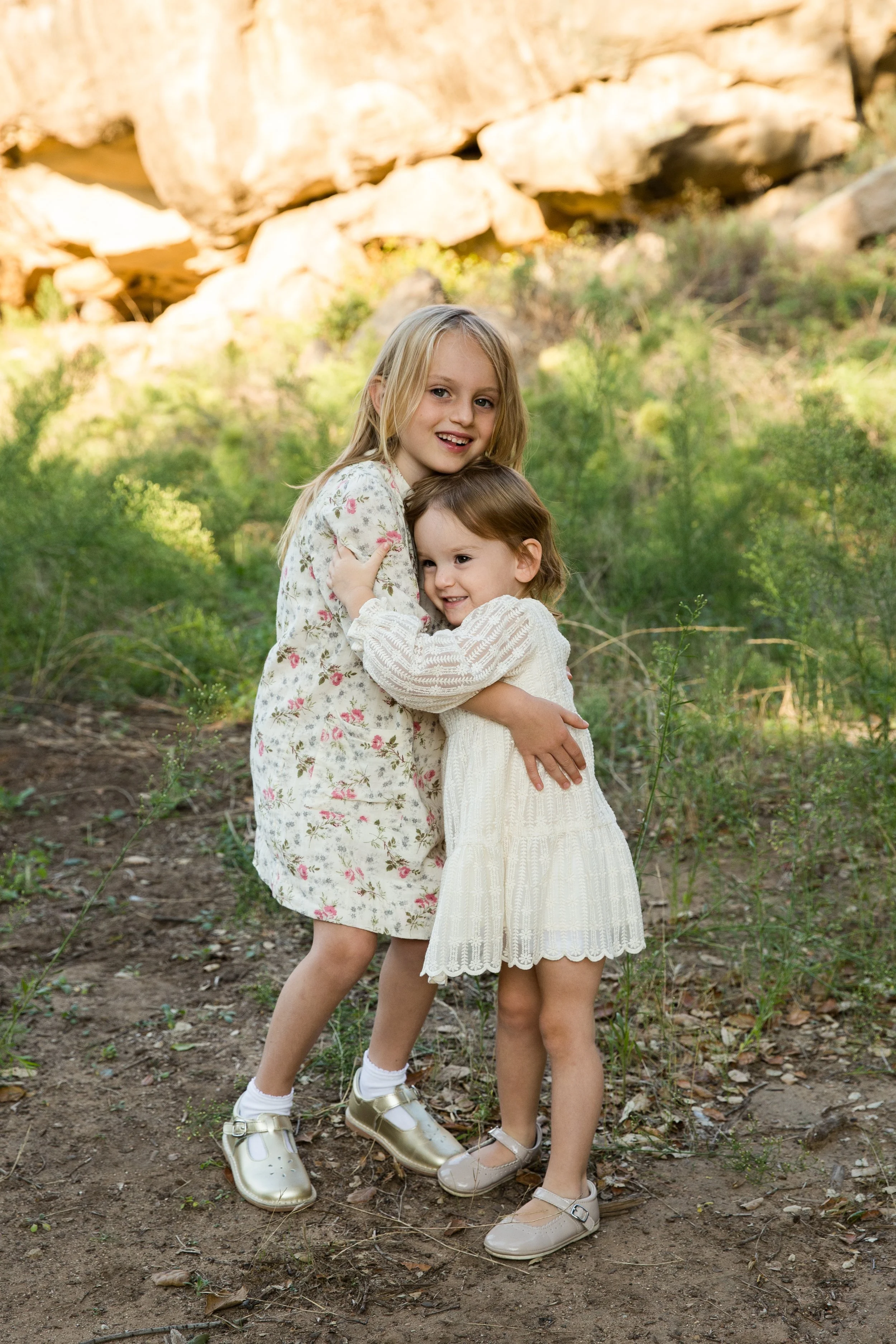 Two young girls are hugging outdoors in a natural setting with rocks and greenery in the background, smiling happily.