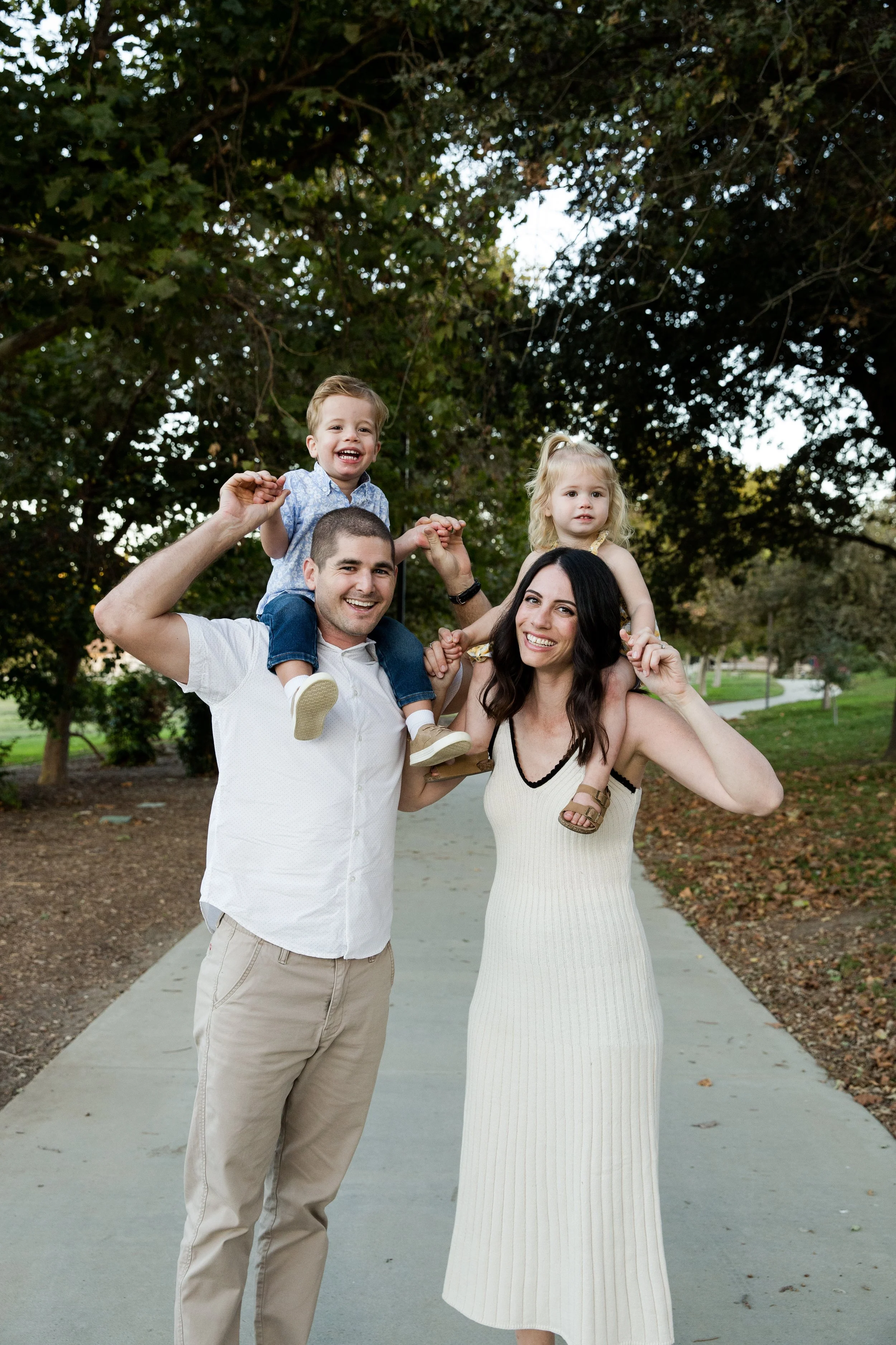 A family of four smiling and playing outside on a path in a park with trees in the background. The father carries a young boy on his shoulders, and the mother holds a young girl on her shoulders.