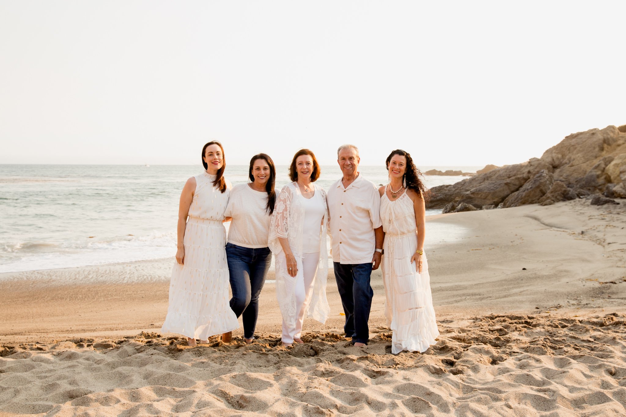 A group of five people, three women and two men, standing on a beach during sunset, all dressed in white and smiling.