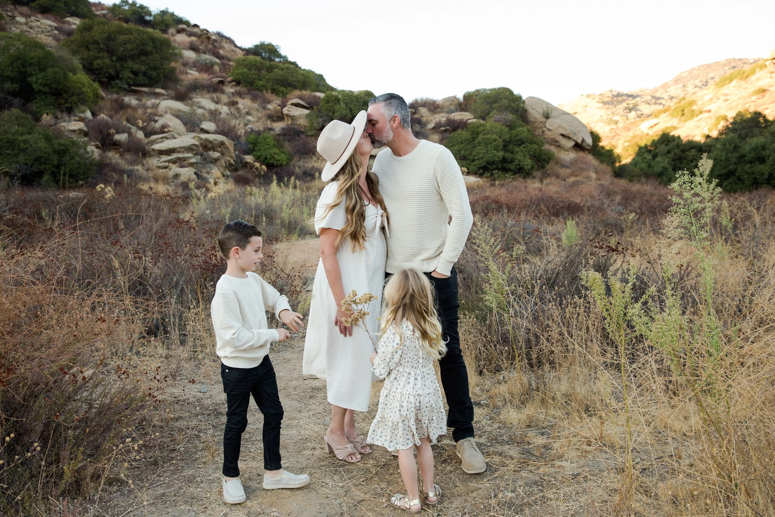 Family of four standing on a dirt trail in a dry, grassy landscape with rocky hills in the background. The parents are kissing, with the mother wearing a wide-brimmed hat and the father in a cream sweater. The two children, a boy and a girl, are near