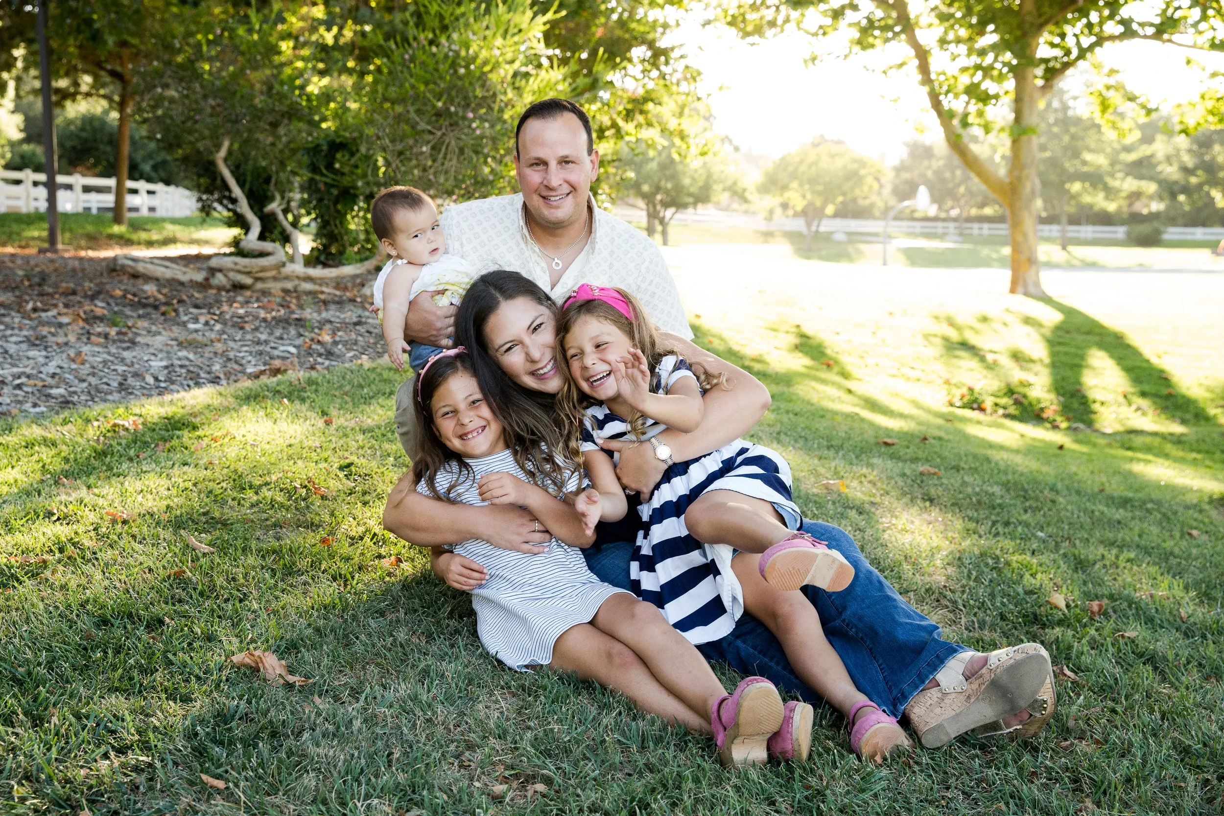 A happy family of five sitting on the grass in a park, smiling and hugging each other with trees and a fountain in the background.