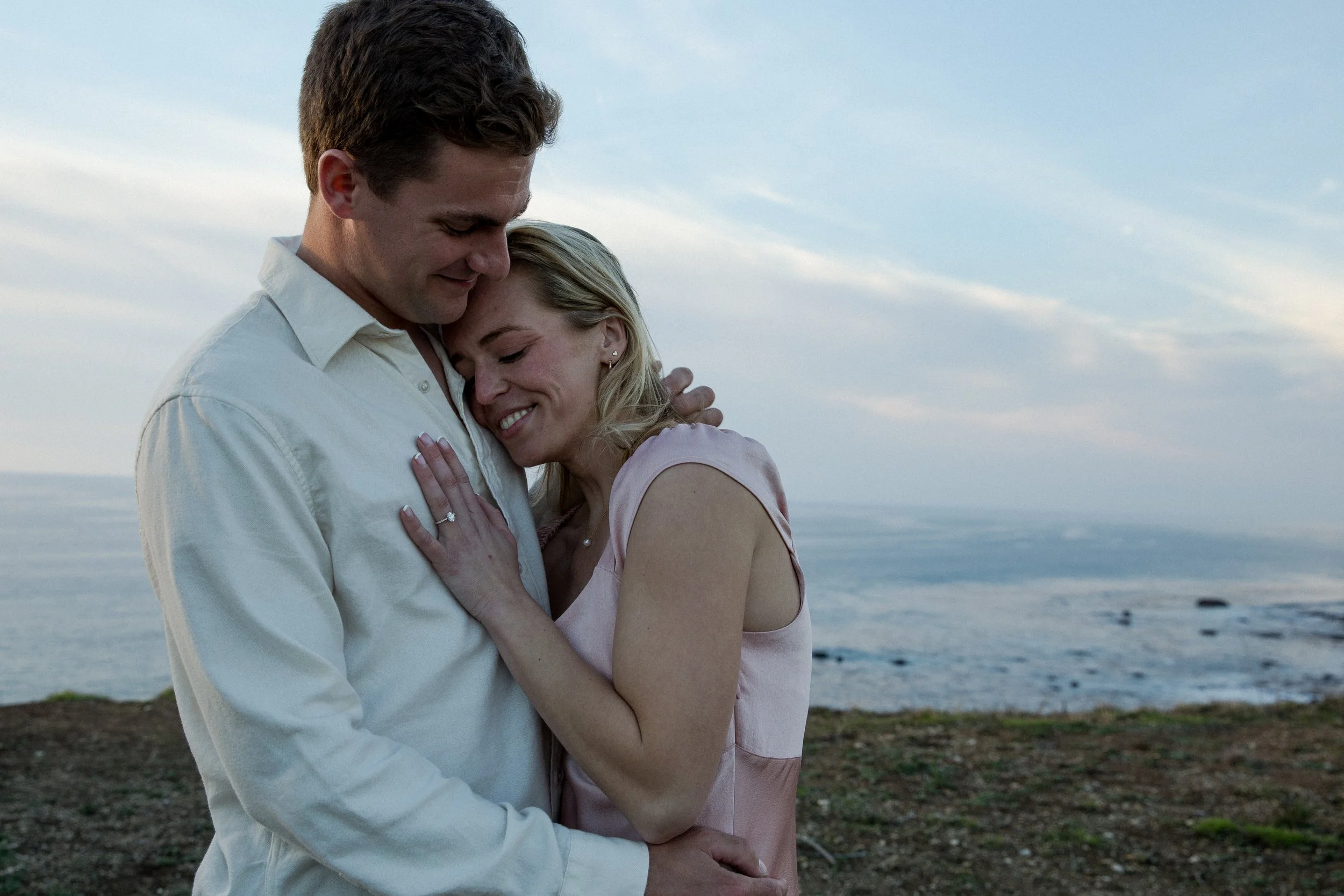 A couple embracing on a beach during sunset, with the ocean and sky in the background.
