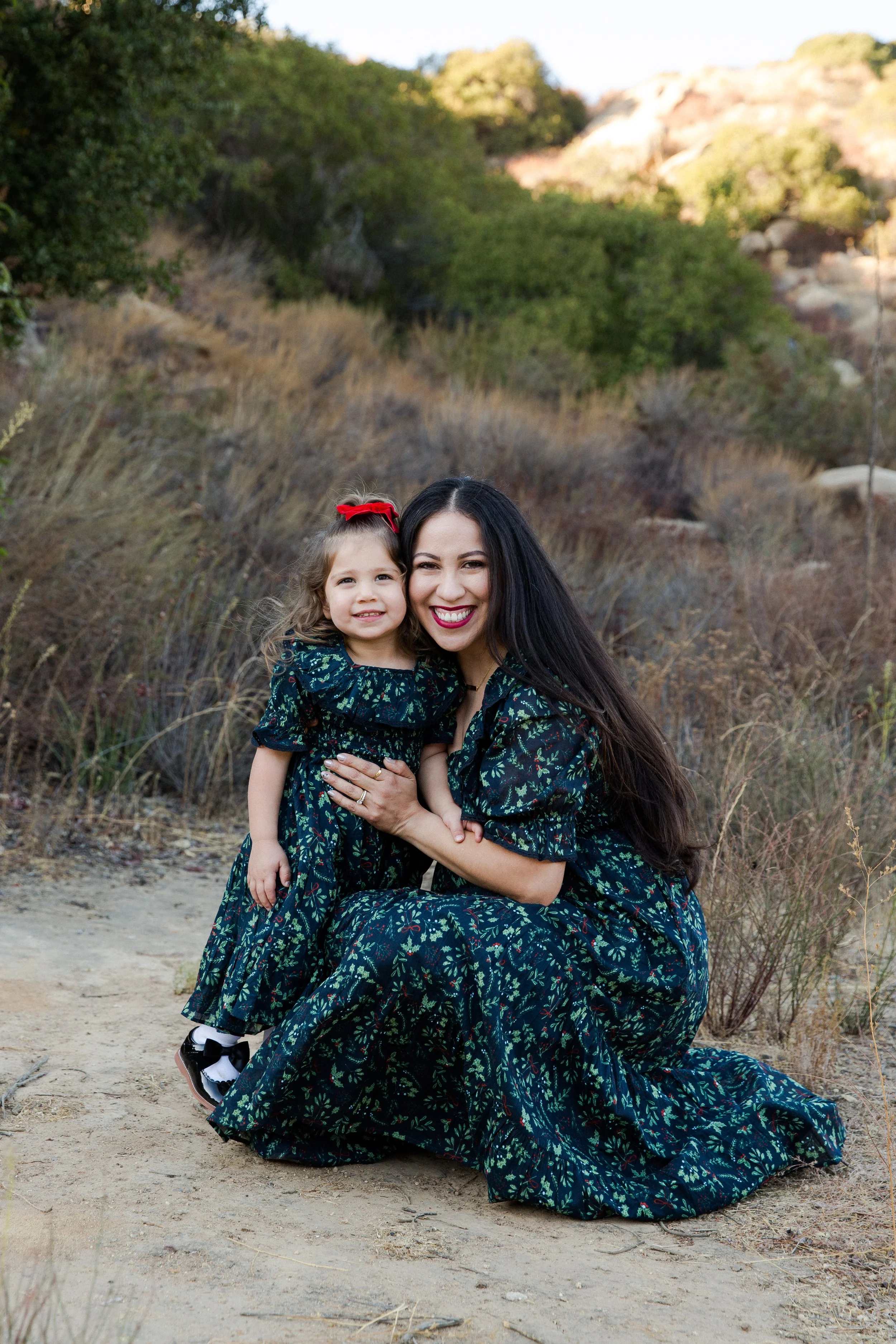 A woman and a young girl, both dressed in matching dark floral dresses, are outdoors in a natural setting with dry grass and bushes. The woman has long dark hair and is smiling, kneeling on the ground while holding the girl, who has curly hair tied w