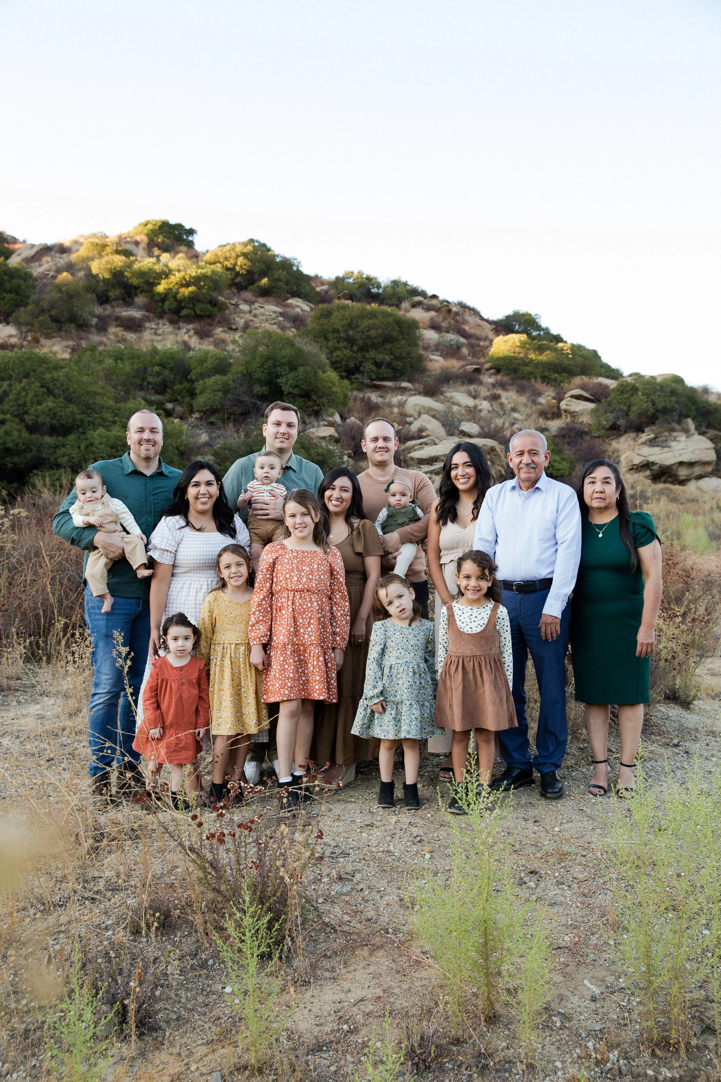 Family group photo outdoors with adults and children, standing on dry ground in front of a hill with sparse vegetation.