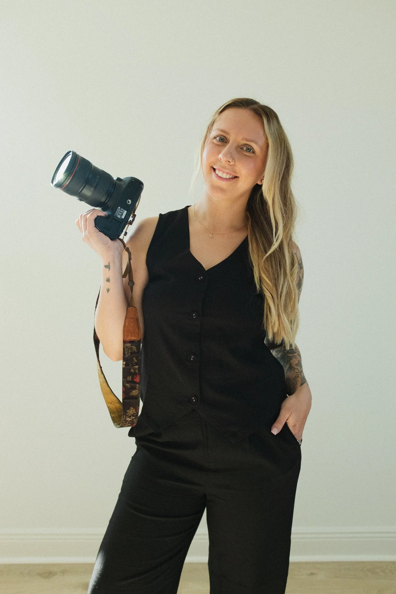 A woman with long blonde hair, wearing a black sleeveless top and black pants, holds a camera in her right hand and smiles at the camera.