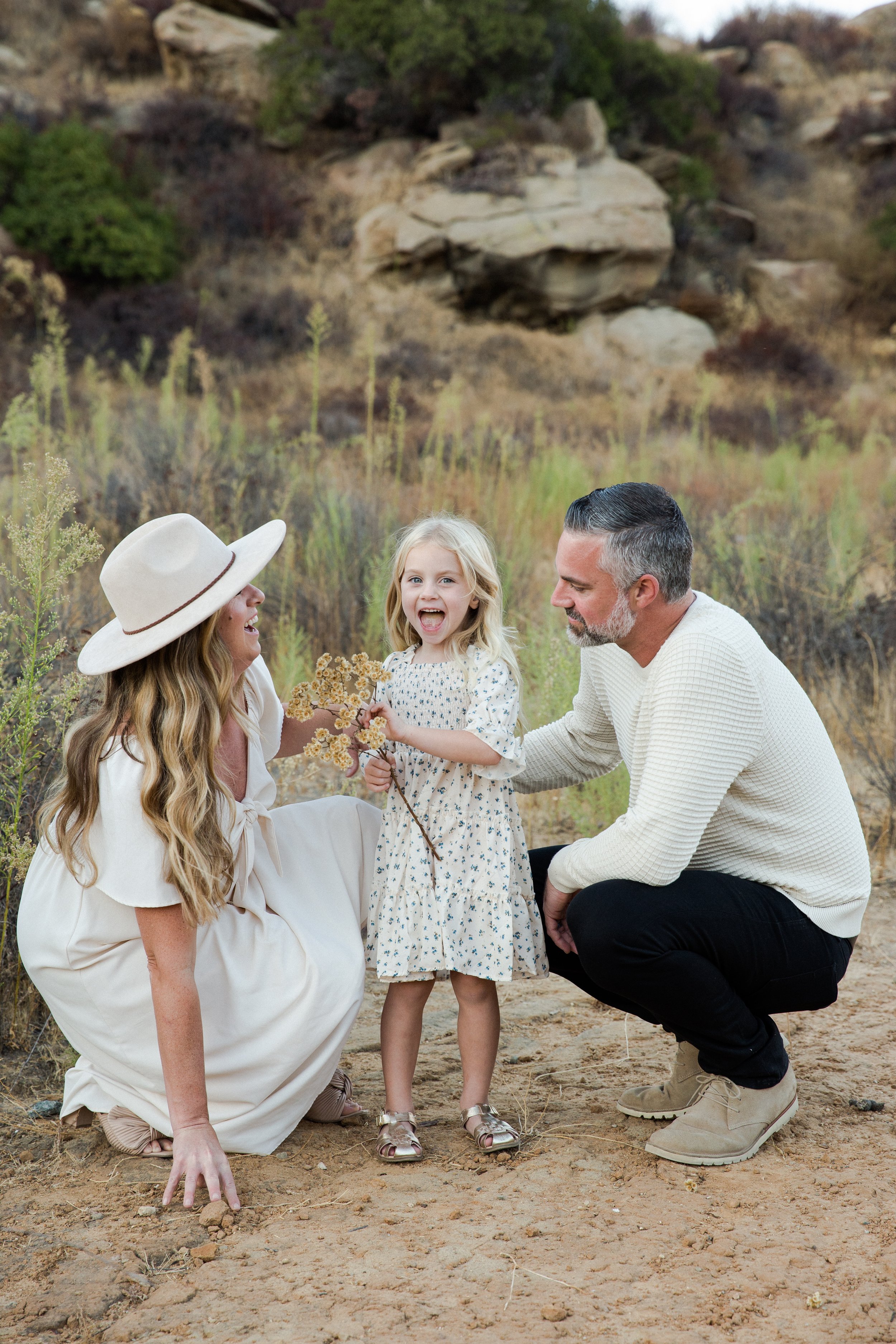 A family of three with a girl, sitting on dirt outdoors in a desert landscape, smiling and interacting with each other.