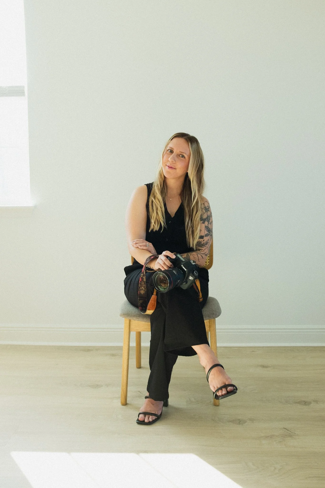 A woman with long blonde hair, wearing a black sleeveless top and black pants, is sitting on a chair in a bright, minimalist room with white walls and wooden flooring. She has a camera on her lap and has a floral tattoo on her left arm, with a slightly puzzled or curious expression on her face.