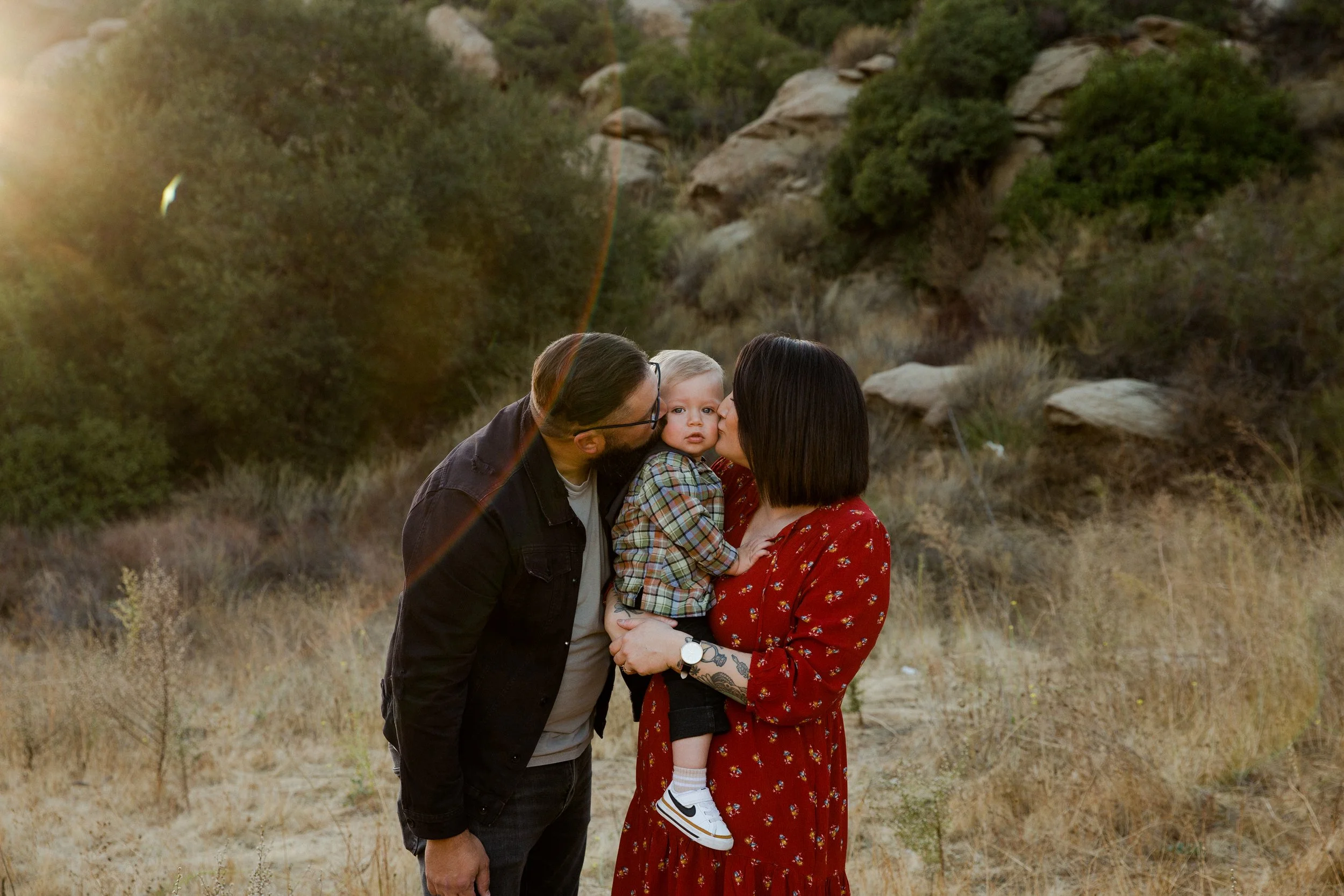 A family of three with a man, woman, and young child outdoors during sunset. The woman is holding the child, and the man is kissing the child's cheek. The background features dry grass, rocks, and bushes.