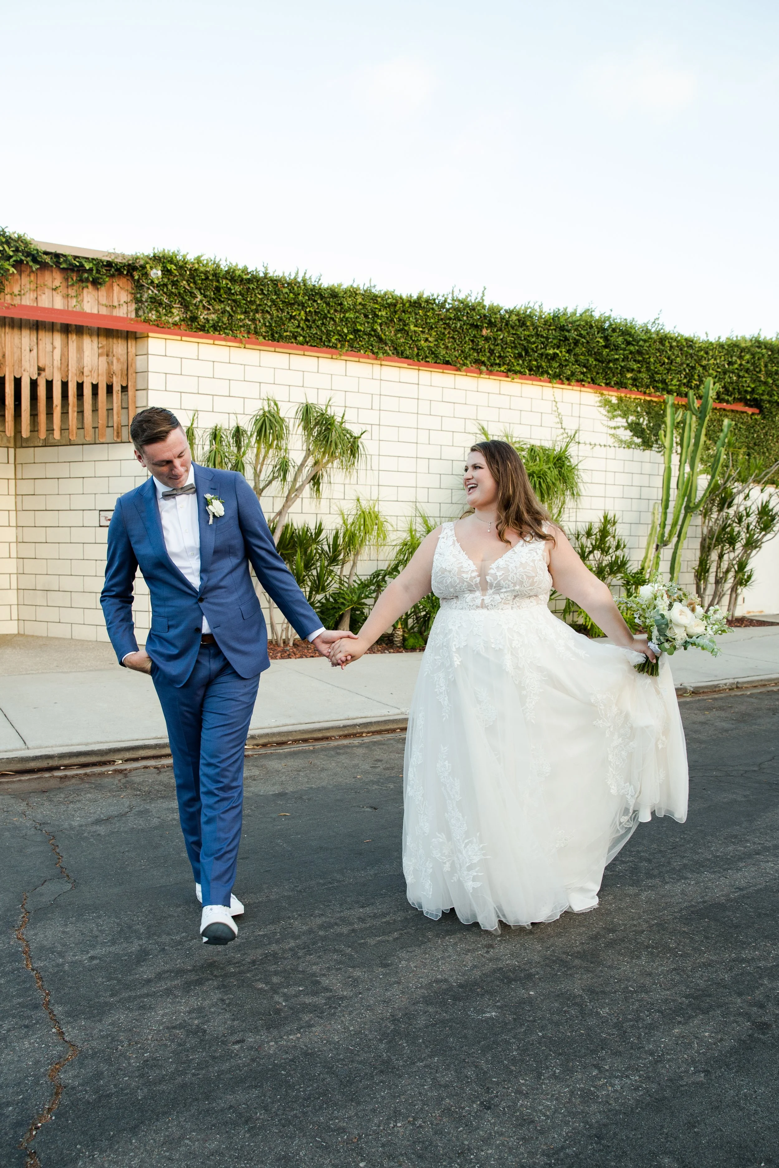A bride and groom holding hands and smiling while walking outside on a paved street in a wedding dress and suit.