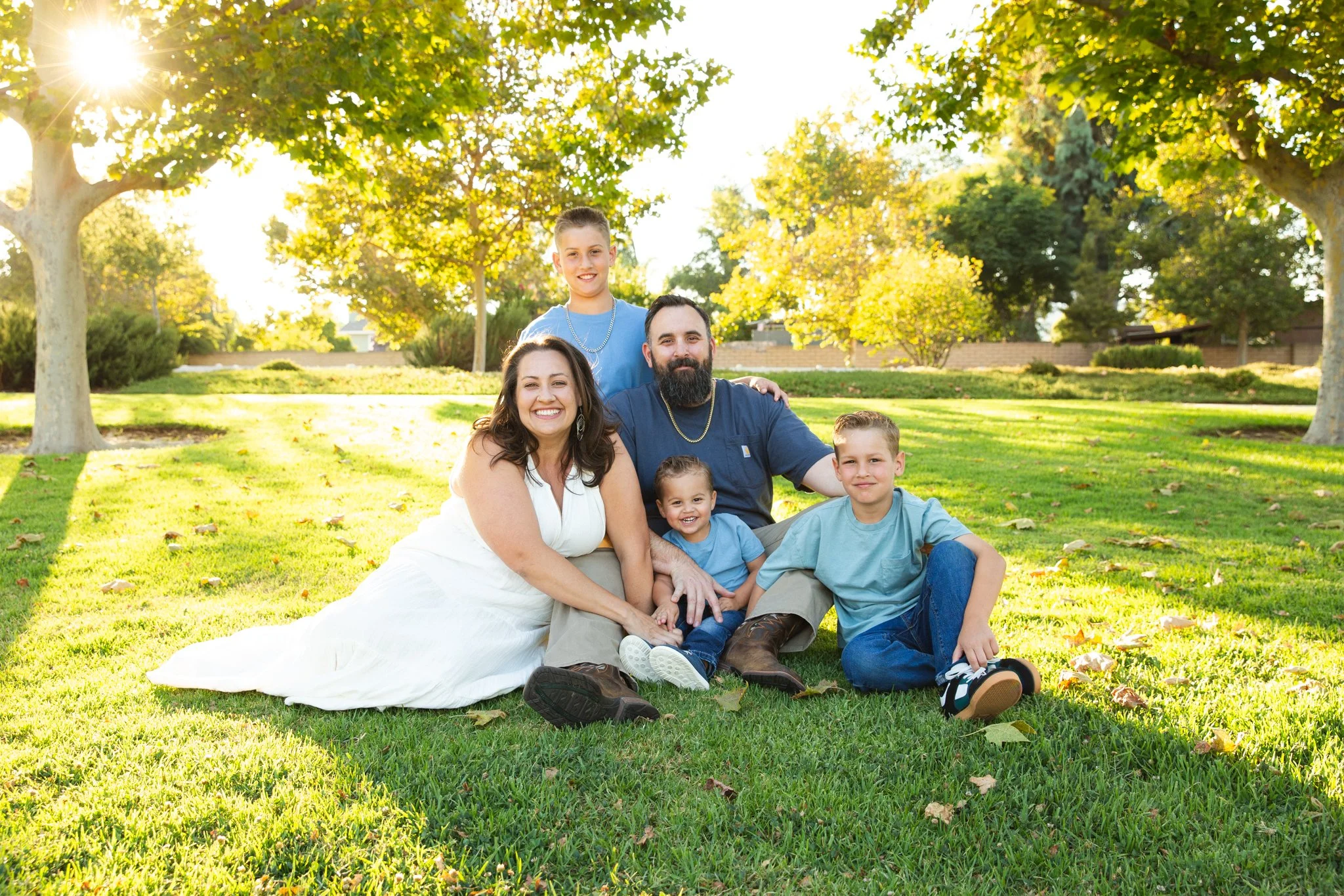 A happy family of six sitting on the grass in a park during sunset. The family includes a woman in a white dress, a man with a beard, and four children. The park is filled with green trees and autumn leaves on the ground.