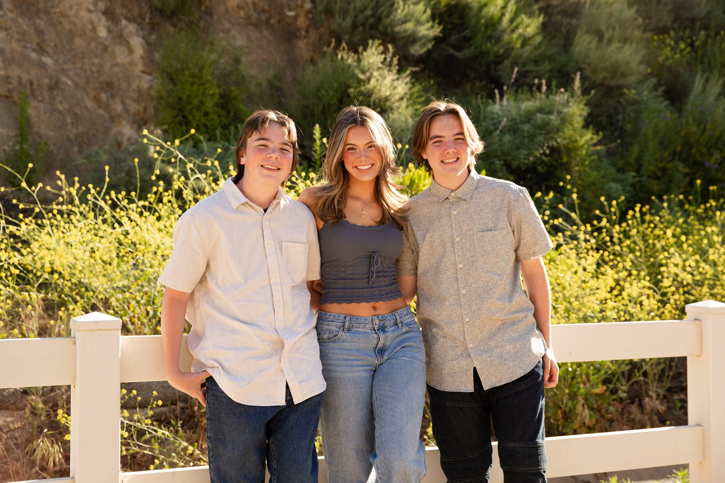 Three teenagers standing outdoors, smiling, with greenery and bushes in the background.
