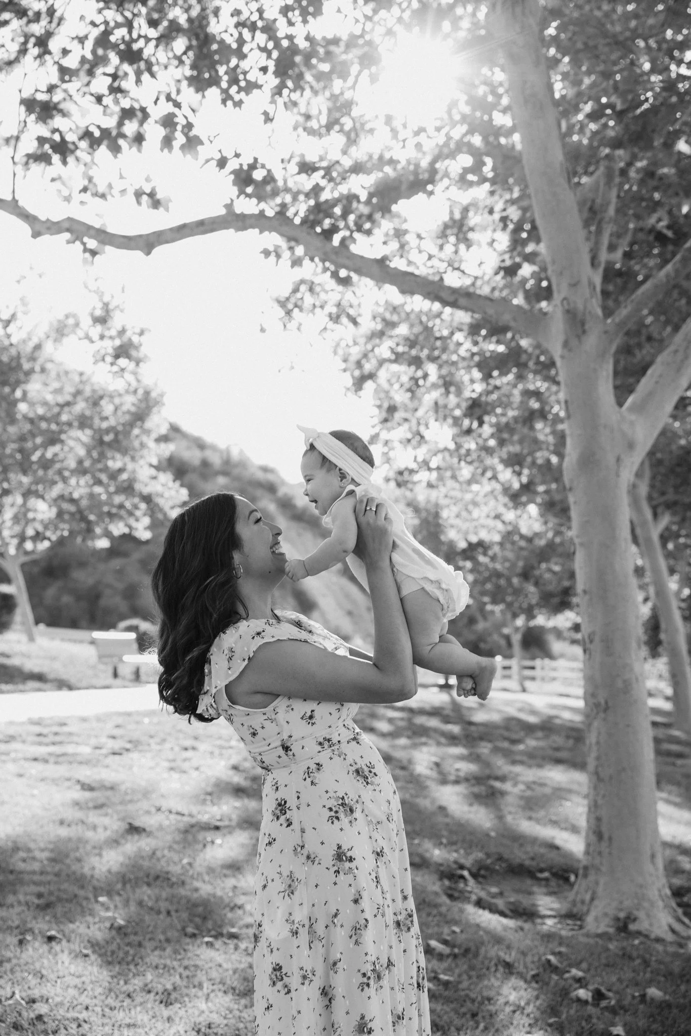 A woman holding a smiling baby girl outdoors in a park with trees and sunlight in the background.