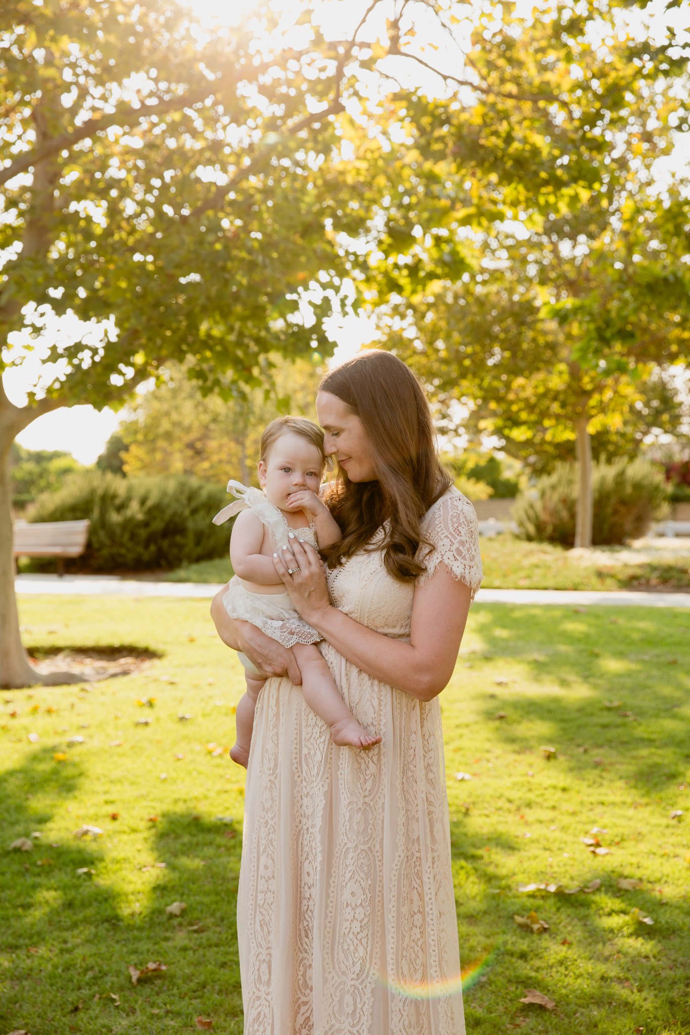 A woman holding a young girl outdoors in a park during sunset, surrounded by trees with green and yellow leaves.