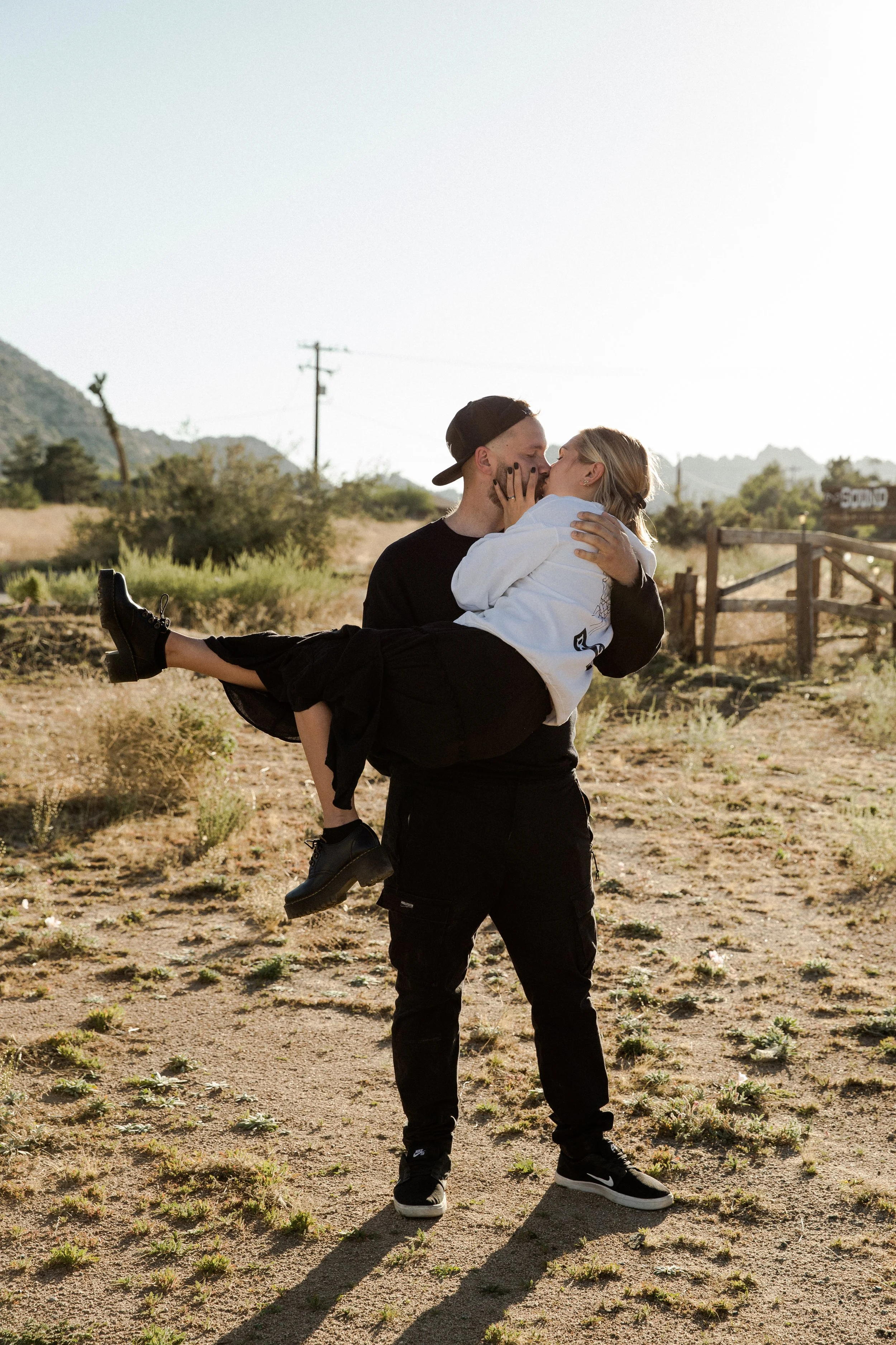 A man in black holding a woman in white in a desert landscape with mountains and power lines in the background.