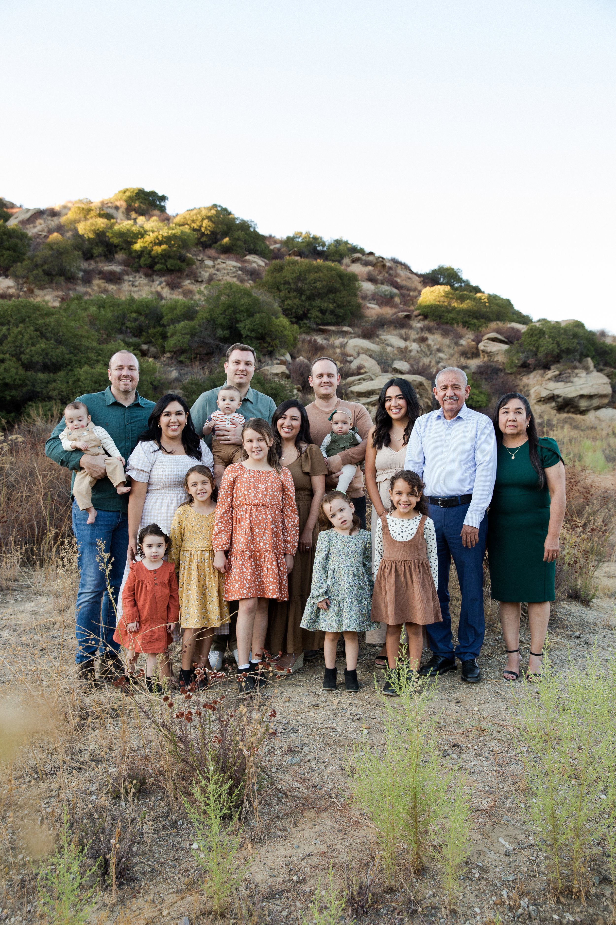 Family photo of adults and children outdoors on a dirt trail with a rocky hillside and shrubs in the background during daytime.