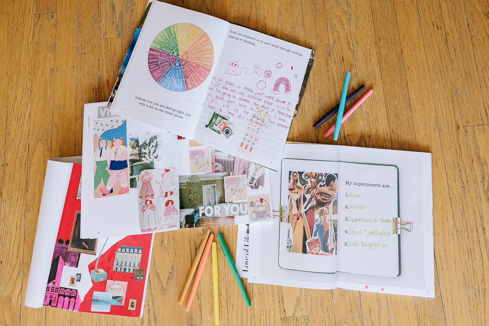 Open magazines and notebooks spread on a wooden surface, with colorful markers nearby, featuring collages, drawings, and lists, including one titled 'My superpowers are...' with five items.