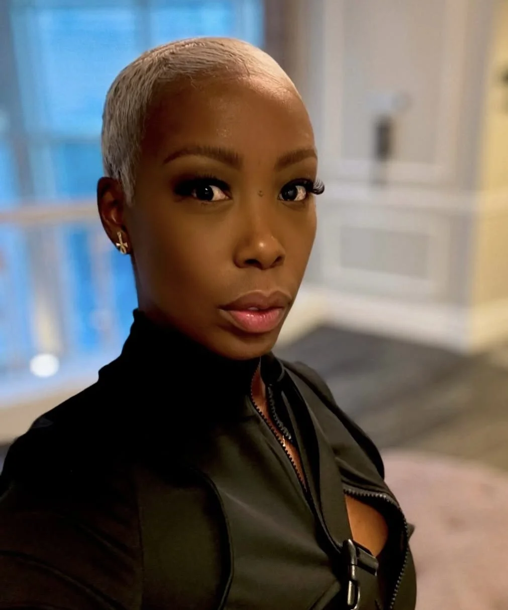 A young woman with short blonde hair, wearing earrings, a black top, and a black zip-up jacket, looking at the camera indoors.