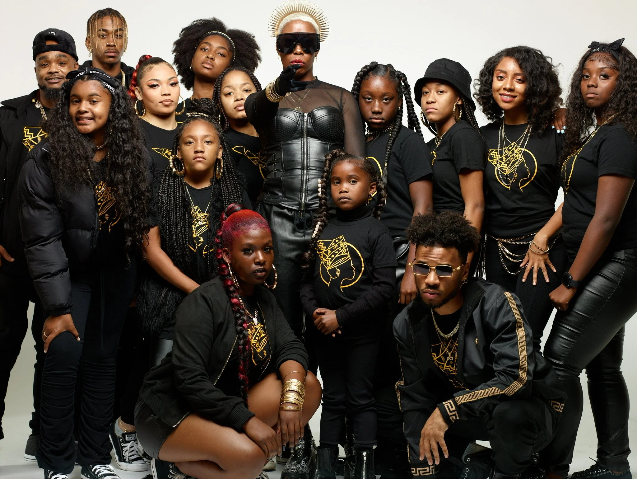 Group of people dressed in black-themed streetwear with gold accents, posing together in front of a white background. One woman in the center wears sunglasses and a halo-style headpiece, with others showing various hairstyles and accessories.