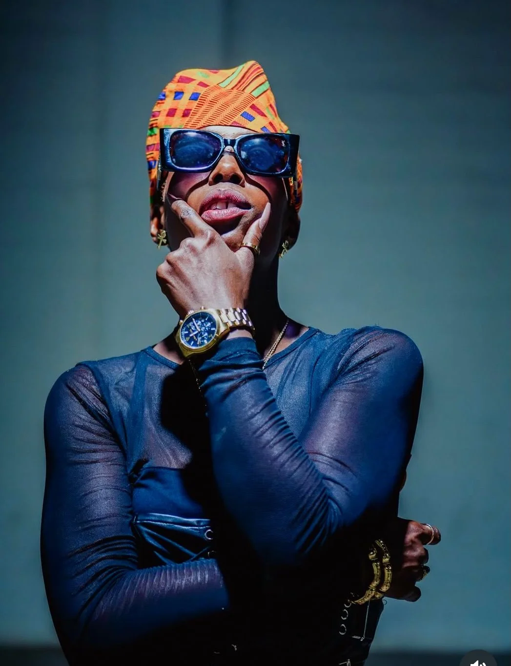A woman wearing large sunglasses, an orange patterned headscarf, and a fitted black top, with a gold watch, rings, and jewelry, posing with her hand on her chin against a plain background.
