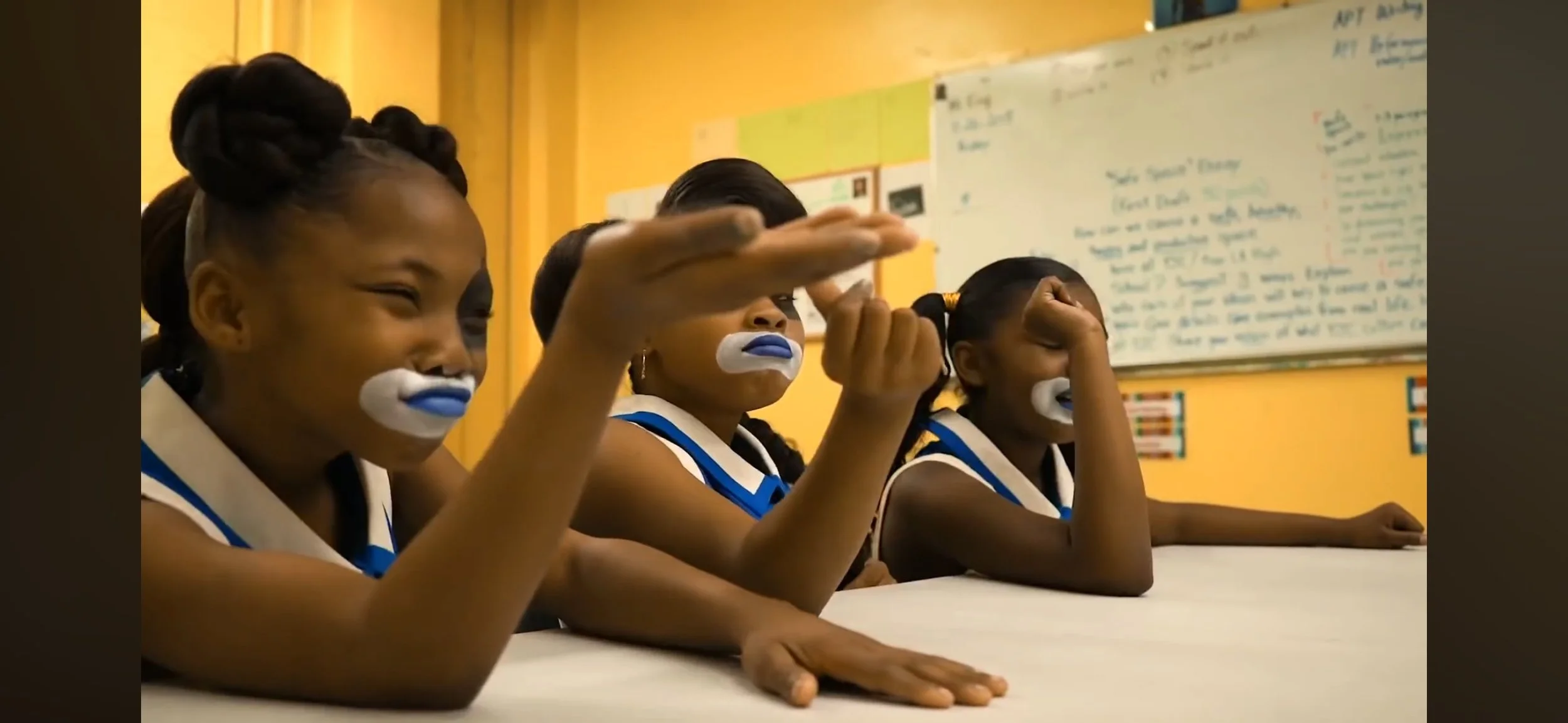Three young girls in a classroom with painted faces, sitting at a table, with a whiteboard with writing in the background.