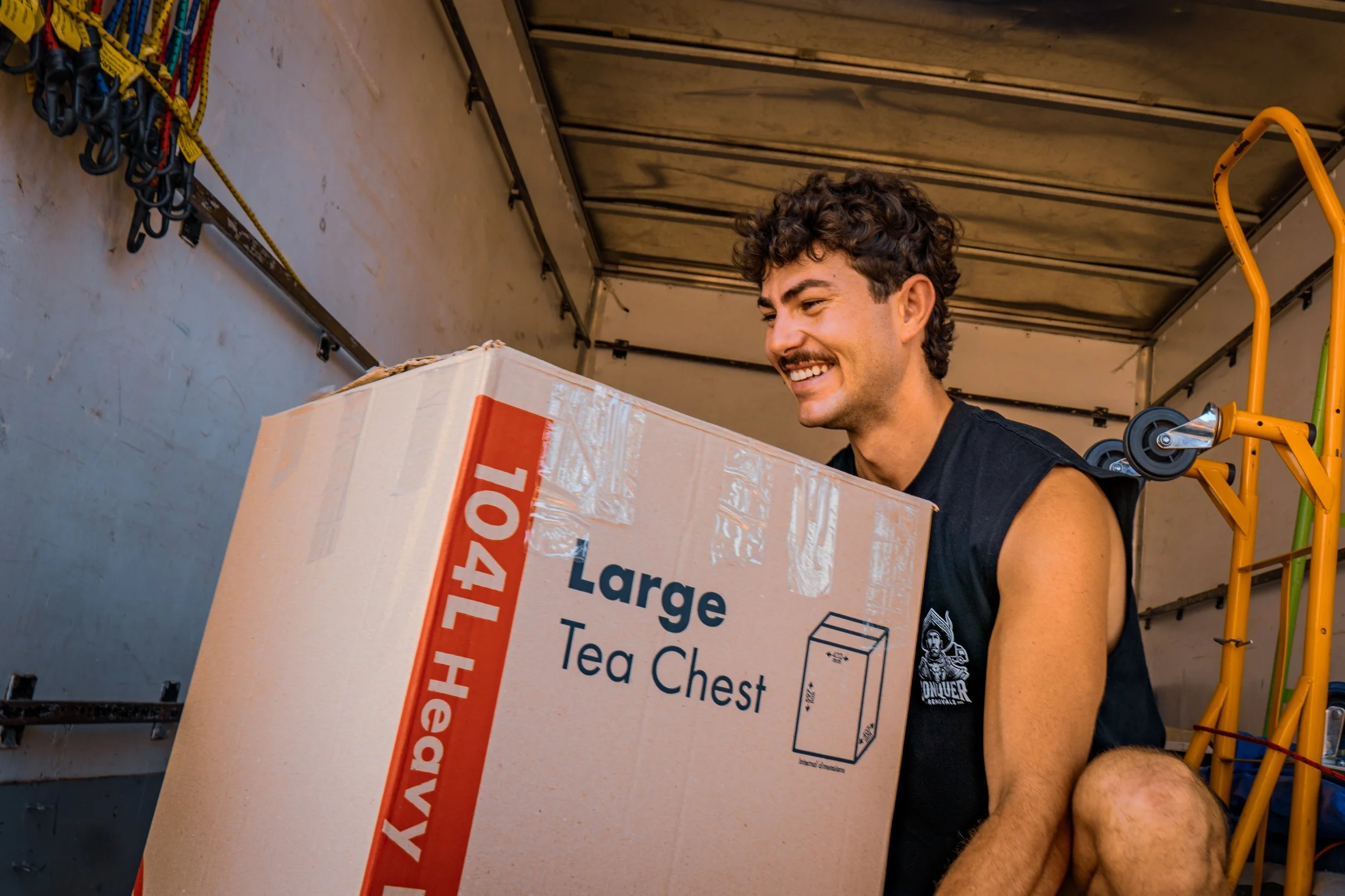 A smiling man loading a large cardboard box labeled 'Large Tea Chest' into a moving truck.