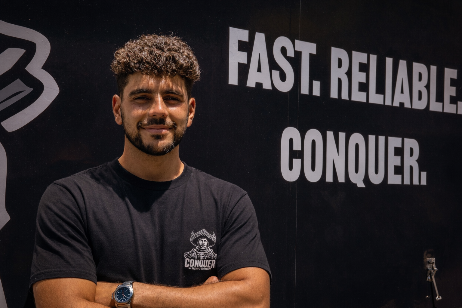 A smiling man with curly hair and a beard, wearing a black t-shirt and a silver watch, standing in front of a dark background with large white text that reads 'Fast. Reliable. Conquer.'