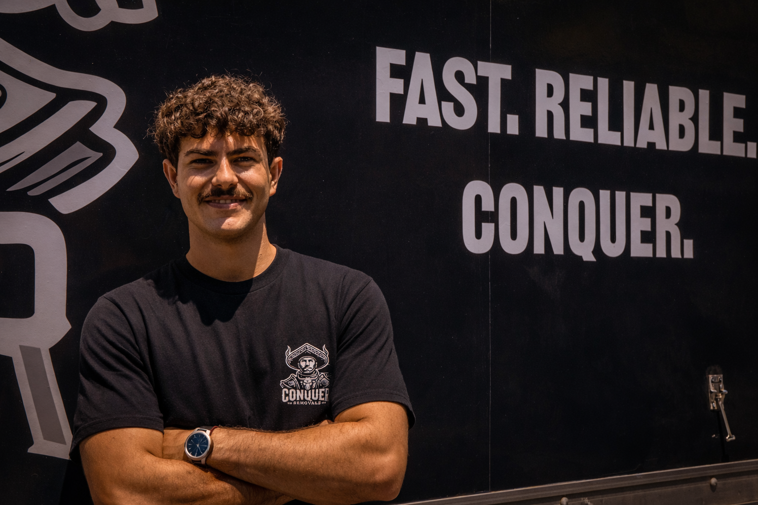 A young man with curly brown hair and a mustache is smiling with arms crossed, standing in front of a black wall with white text that says 'FAST. RELIABLE. CONQUER.'