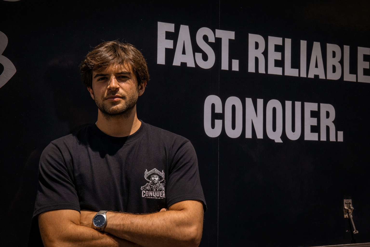 A young man with brown hair and a beard, wearing a black T-shirt with a logo that says 'Conquer,' standing with his arms crossed against a black background with white text that reads, 'Fast. Reliable. Conquer.'