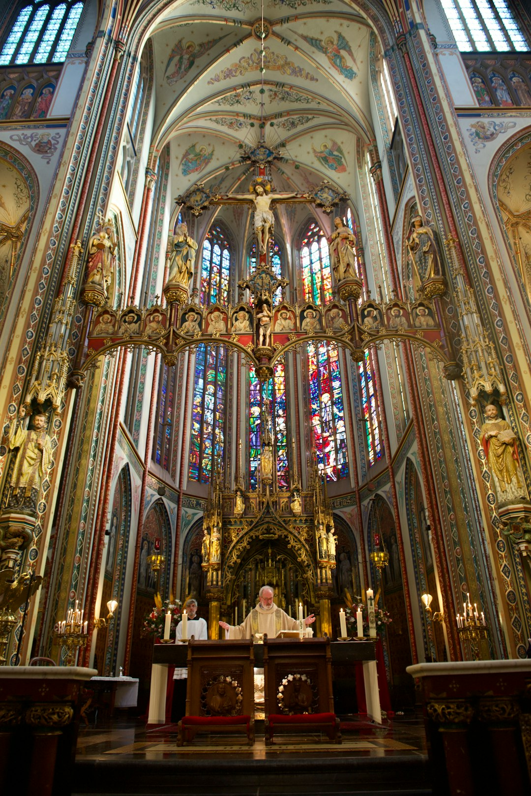 Interior of a colorful church with stained glass windows, ornate gold decorations, statues of saints, and a priest conducting a service at the altar.