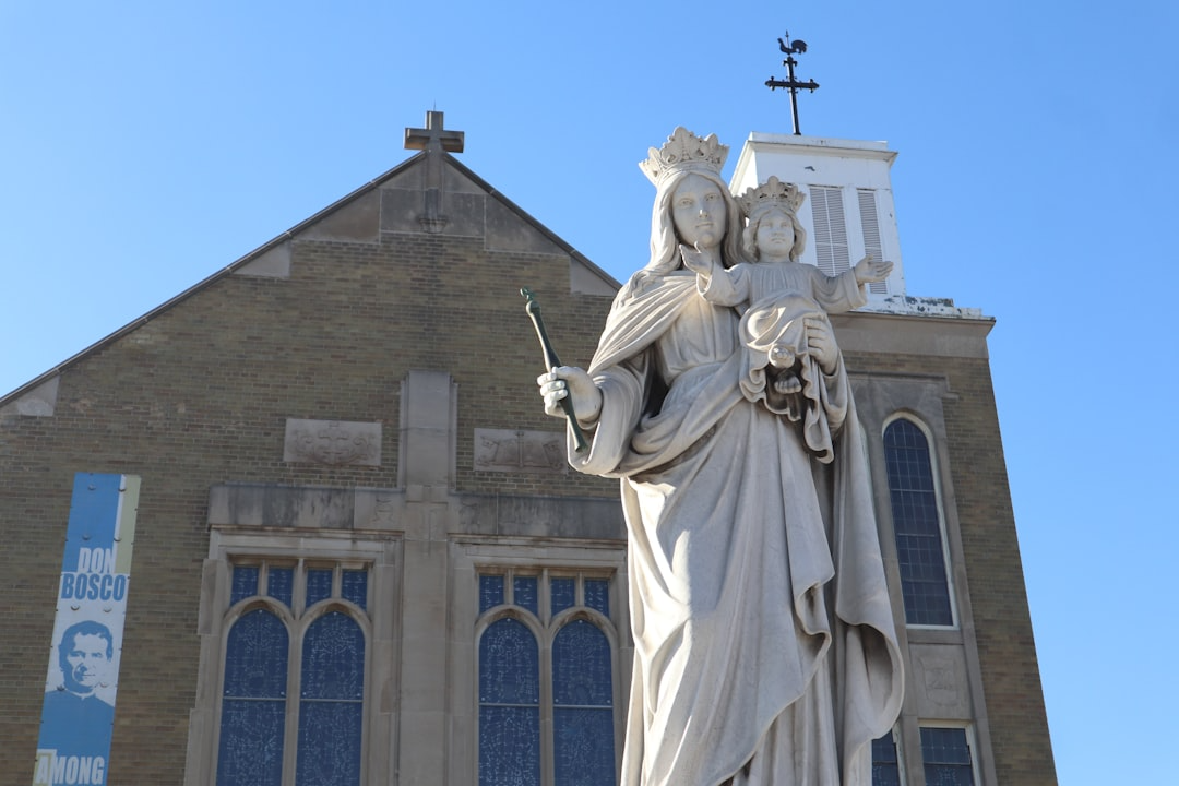 Statue of the Virgin Mary holding baby Jesus in front of a church with a cross on top and blue sky background.